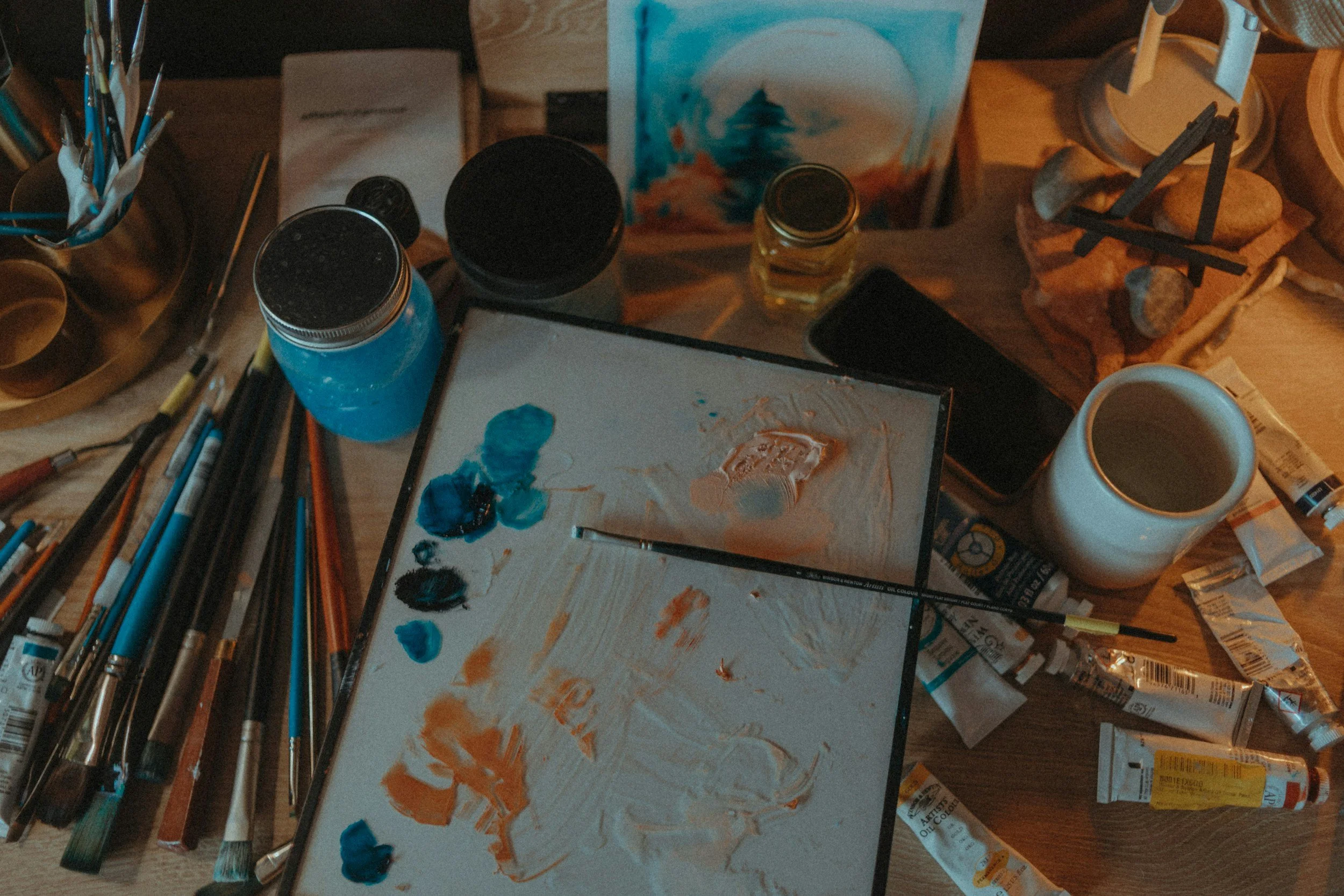 An artist's workspace cluttered with paintbrushes, tubes of paint, jars of blue paint, a palette with mixed colors, a cup, a small canvas with a landscape painting, an iPhone, a candle, and various art supplies.