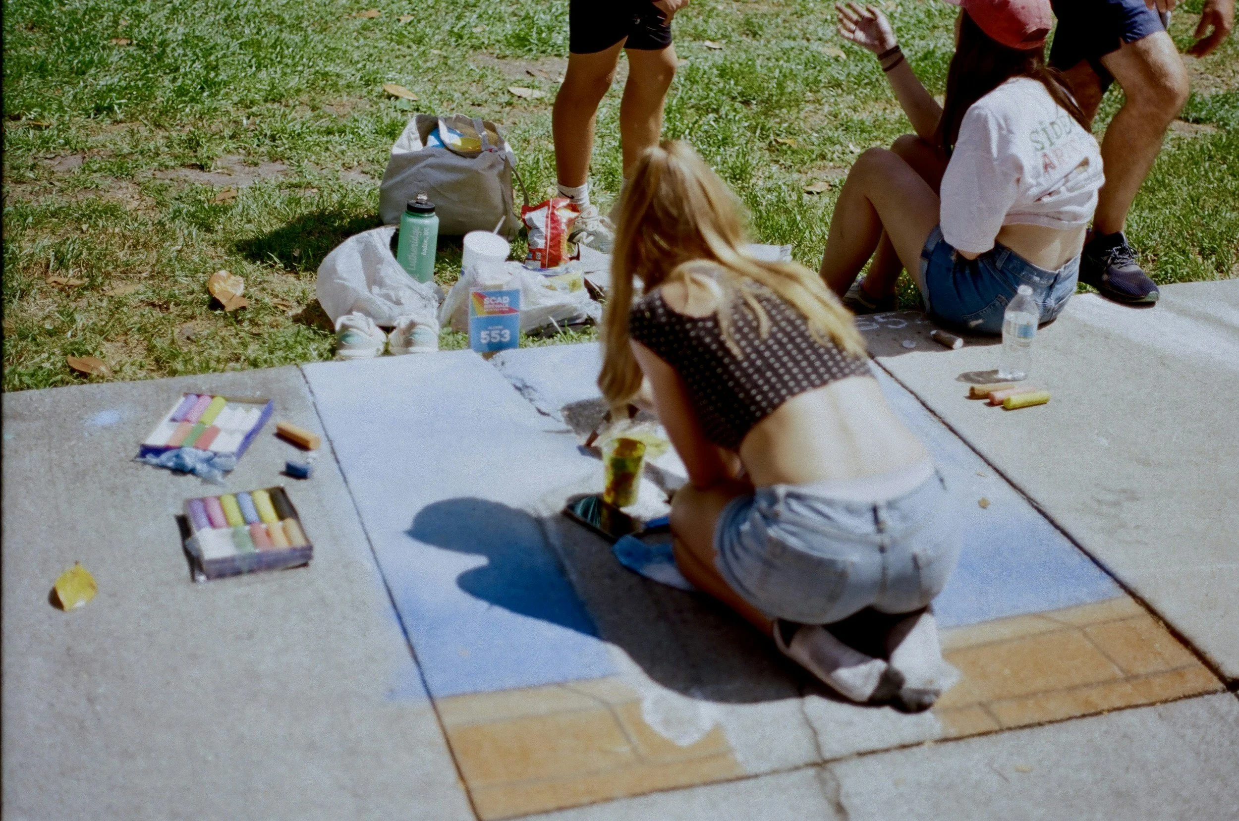 A young woman painting a chalk mural on a sidewalk in a park on a sunny day, surrounded by art supplies and onlookers.