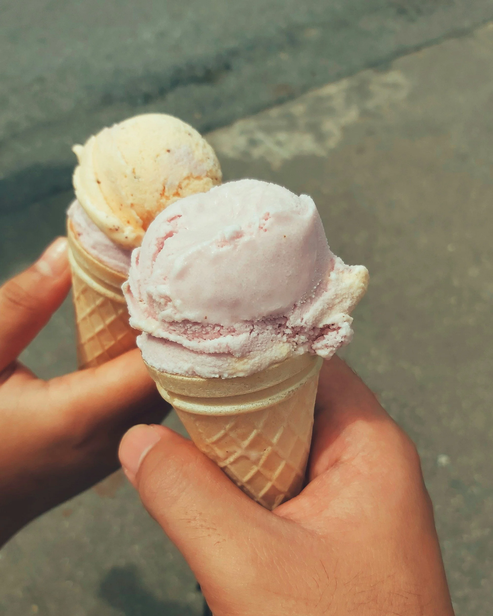 Two hands holding waffle cones with scoops of ice cream on a sidewalk.