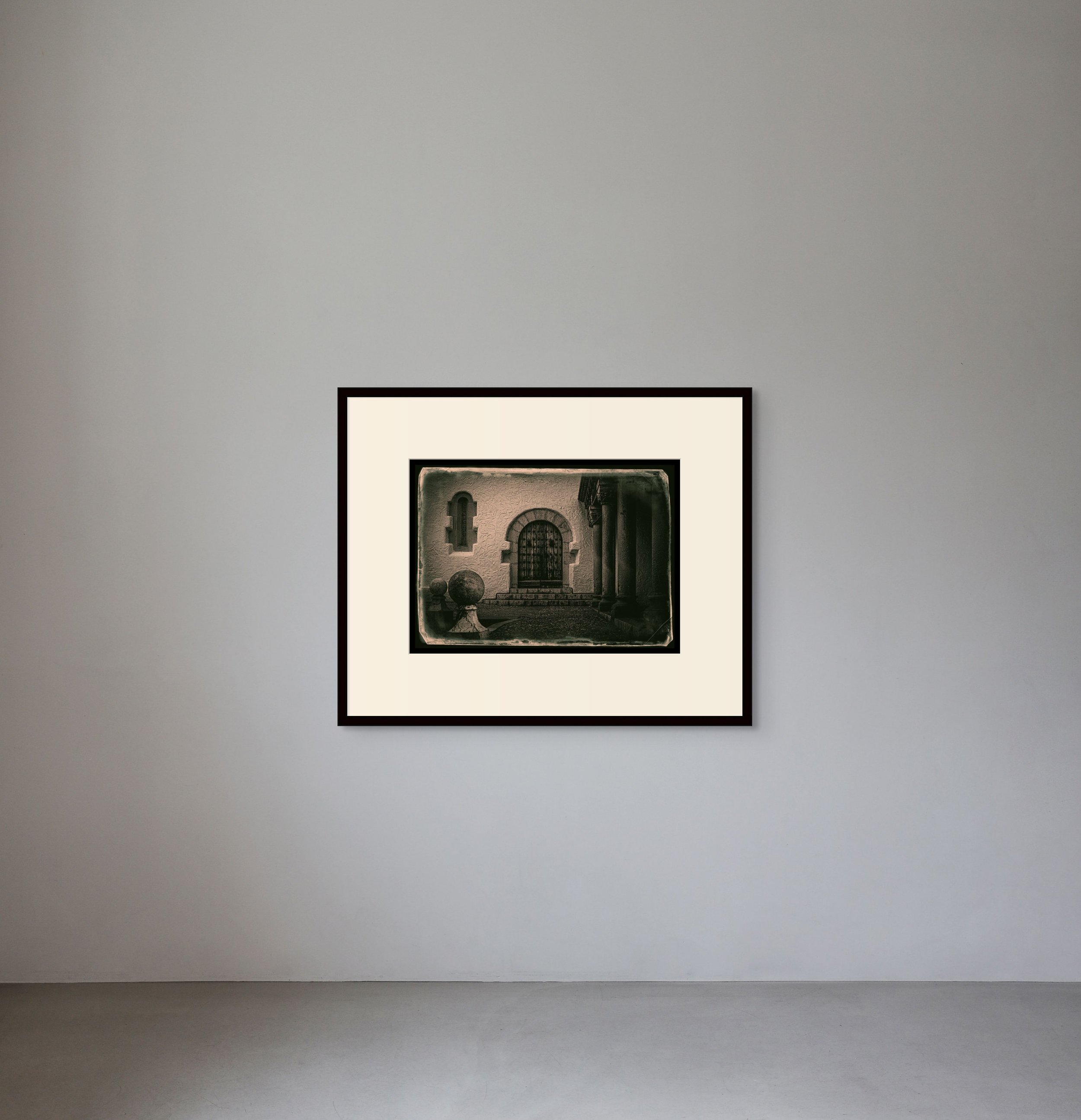 A framed black-and-white photograph of a stone building with a rounded door and small window, hanging on a plain white gallery wall.