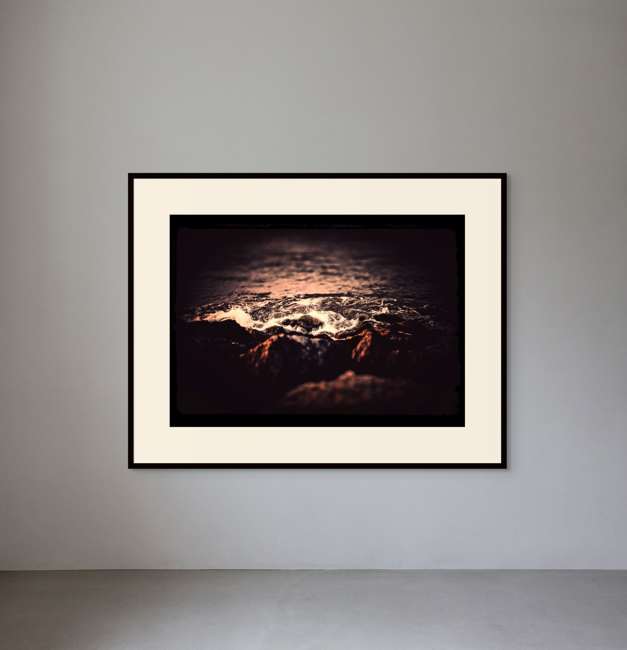 Framed photograph of ocean waves crashing against rocks at sunset, displayed on a gray gallery wall.
