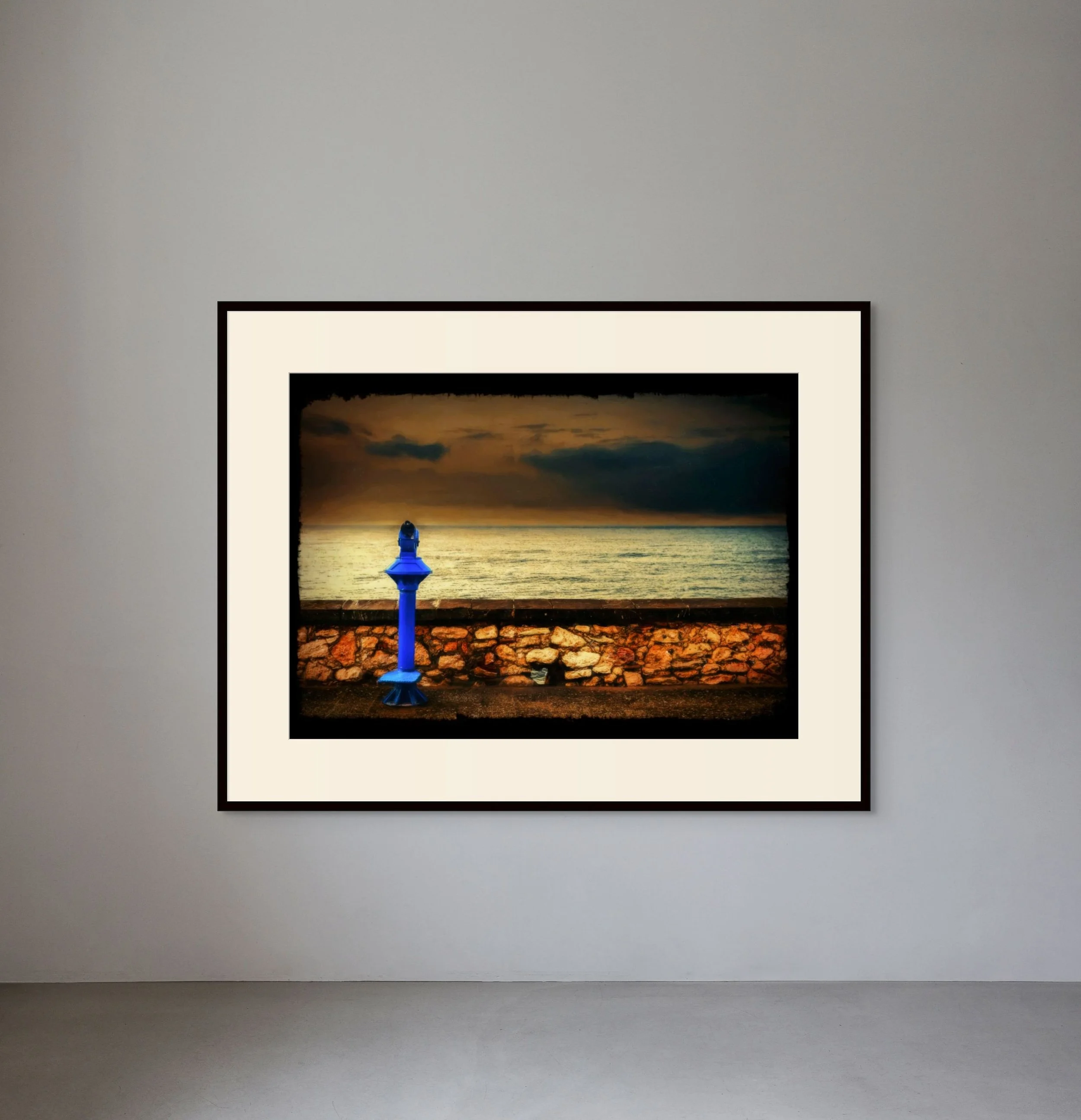 Framed photograph of a sunset over the ocean with a blue lighthouse statue in the foreground and a stone wall along a walkway.