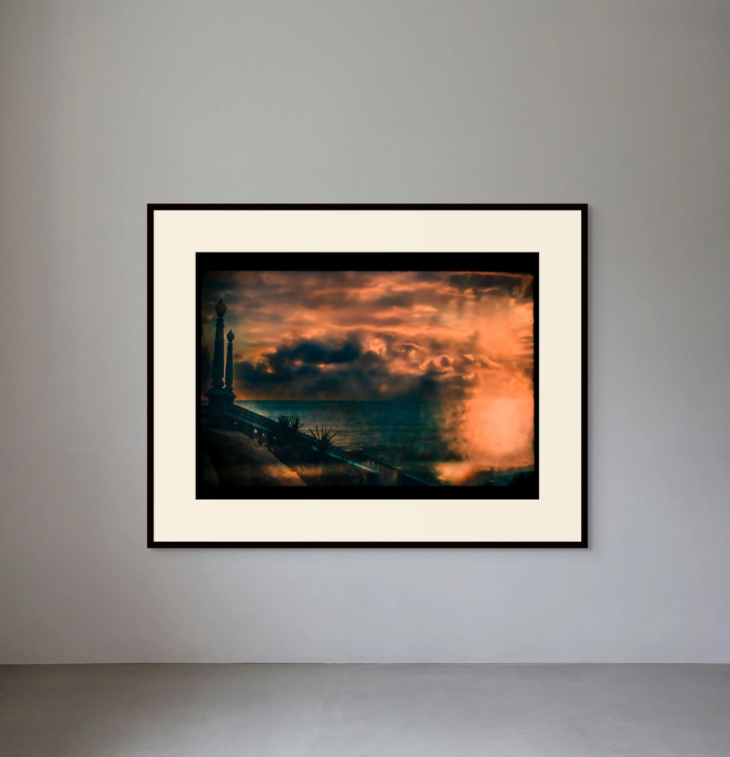 Framed painting of a sunset over the ocean with clouds and a pier with decorative posts along the water's edge