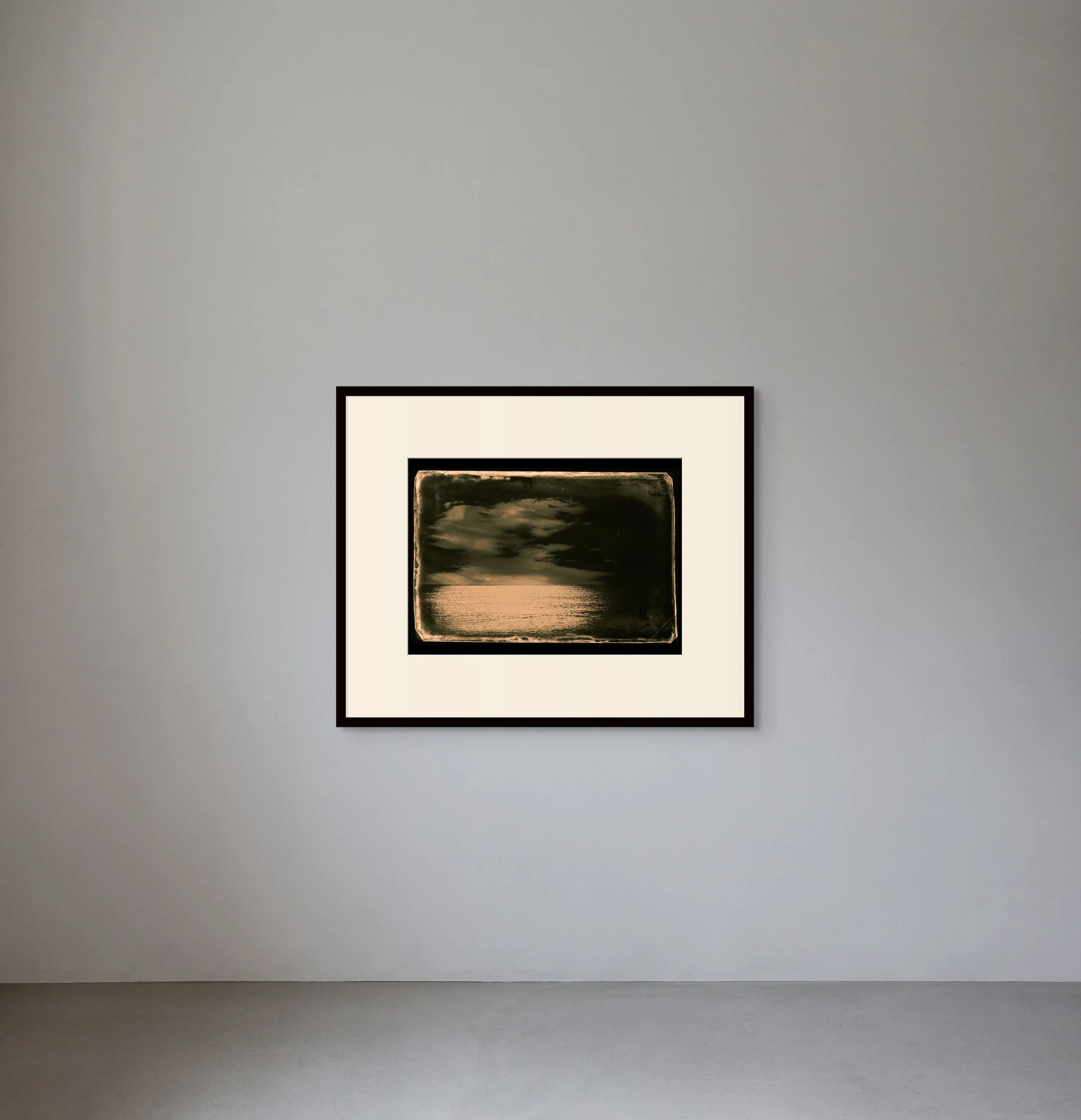 A framed landscape photograph of sky, clouds, and water hangs on a plain white wall in an art gallery.