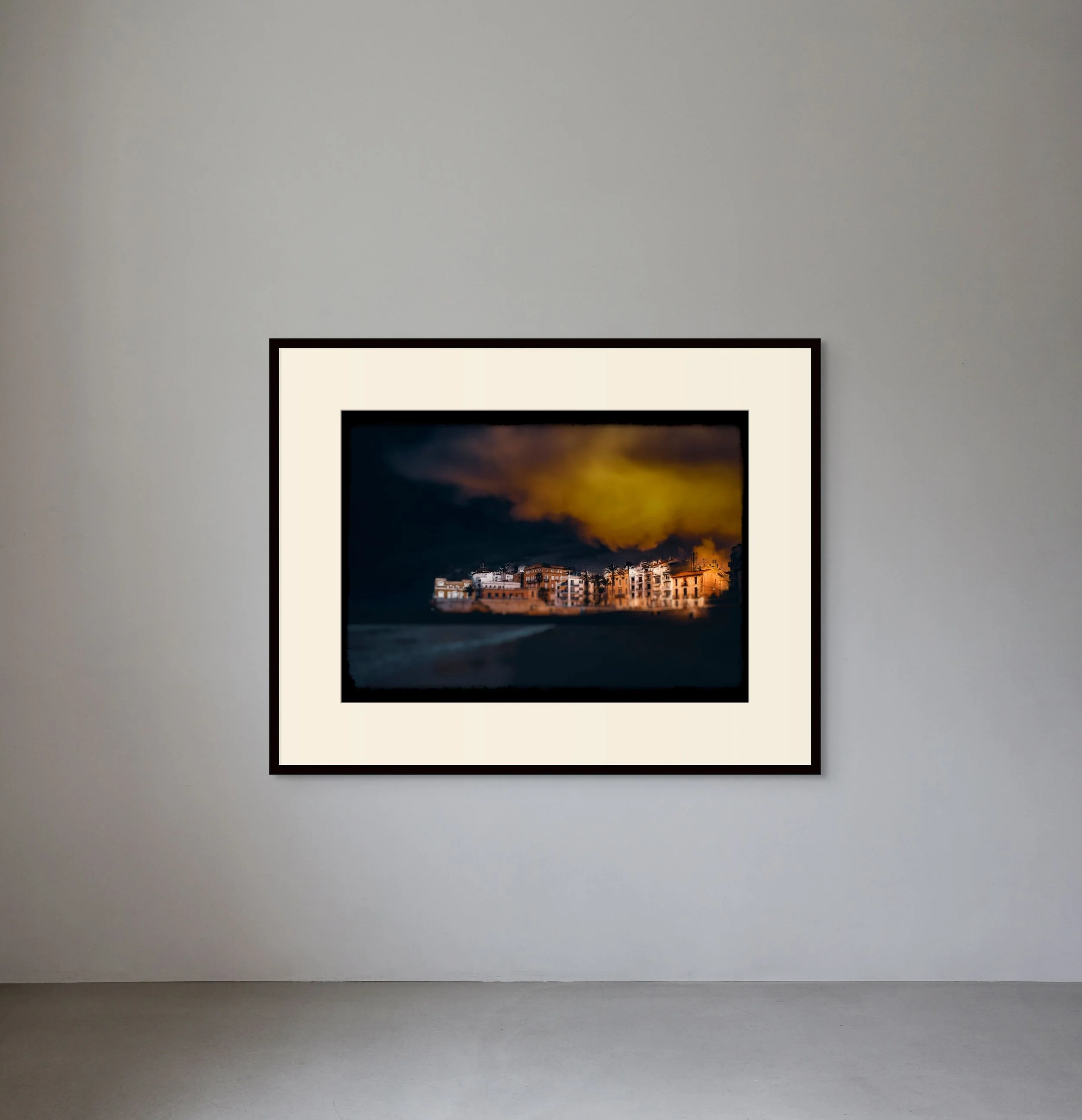 A framed photograph of a coastal town under a dramatic, dark sky with a bright orange glow.