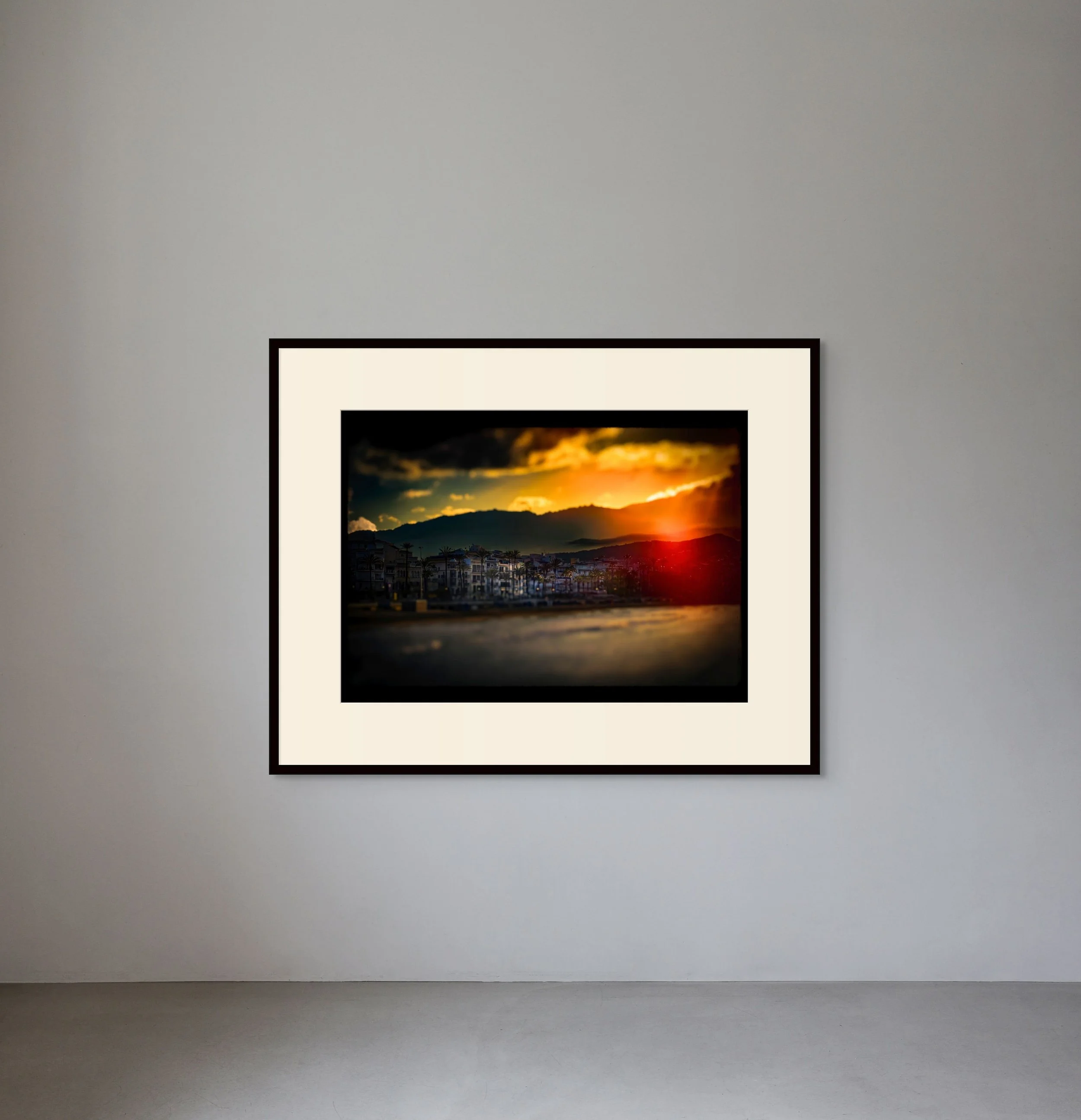 Framed landscape photo of a sunset over mountains and a city with palm trees, hanging on a plain white wall.