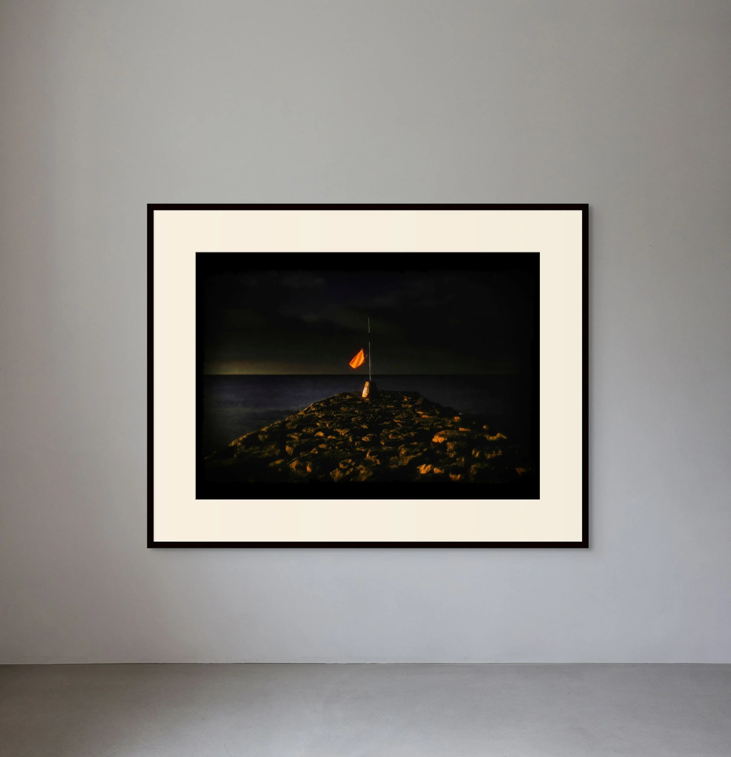 A framed photograph of a rocky breakwater at night, with a small orange lighthouse or beacon at the end, against a dark sky and ocean background.