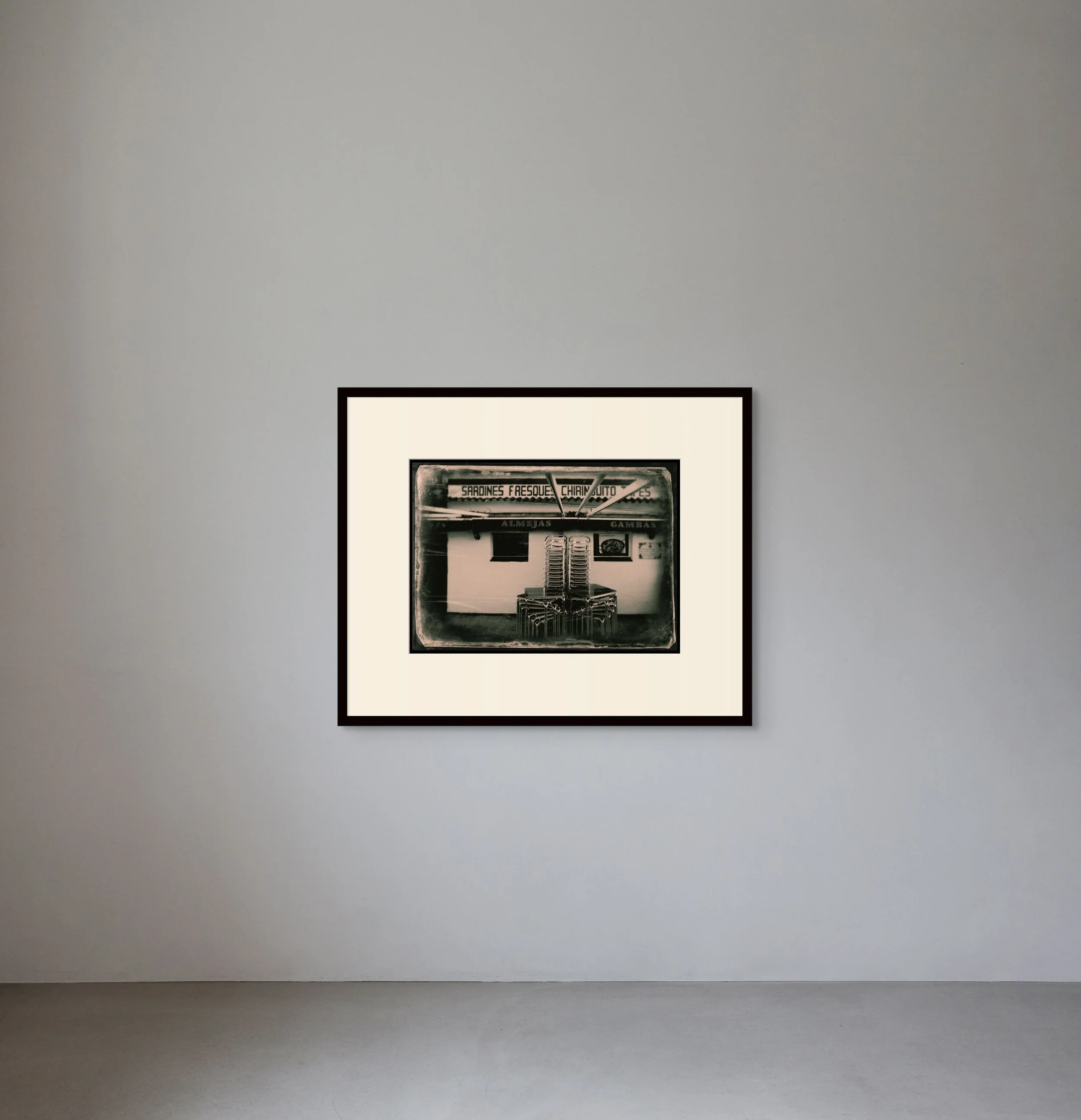 Black and white framed photograph of a vintage kiosk or stand selling canned sardines, with stacked chairs in front and Spanish text on the sign.