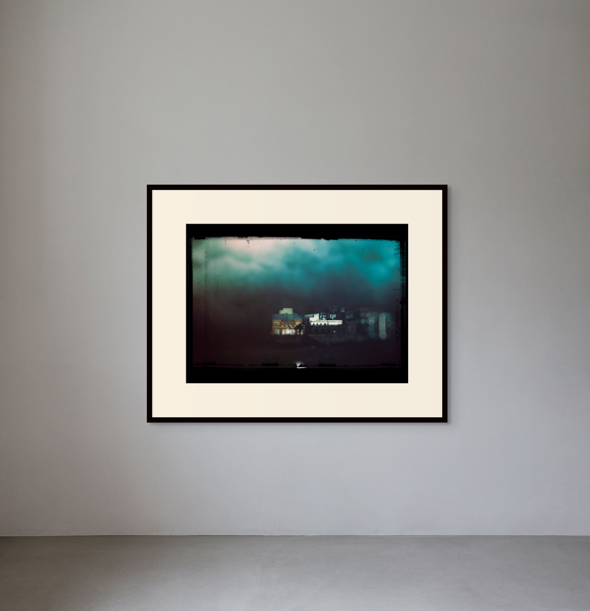 Photograph of a framed abstract artwork on a white gallery wall, depicting a cityscape with dark stormy clouds overhead.