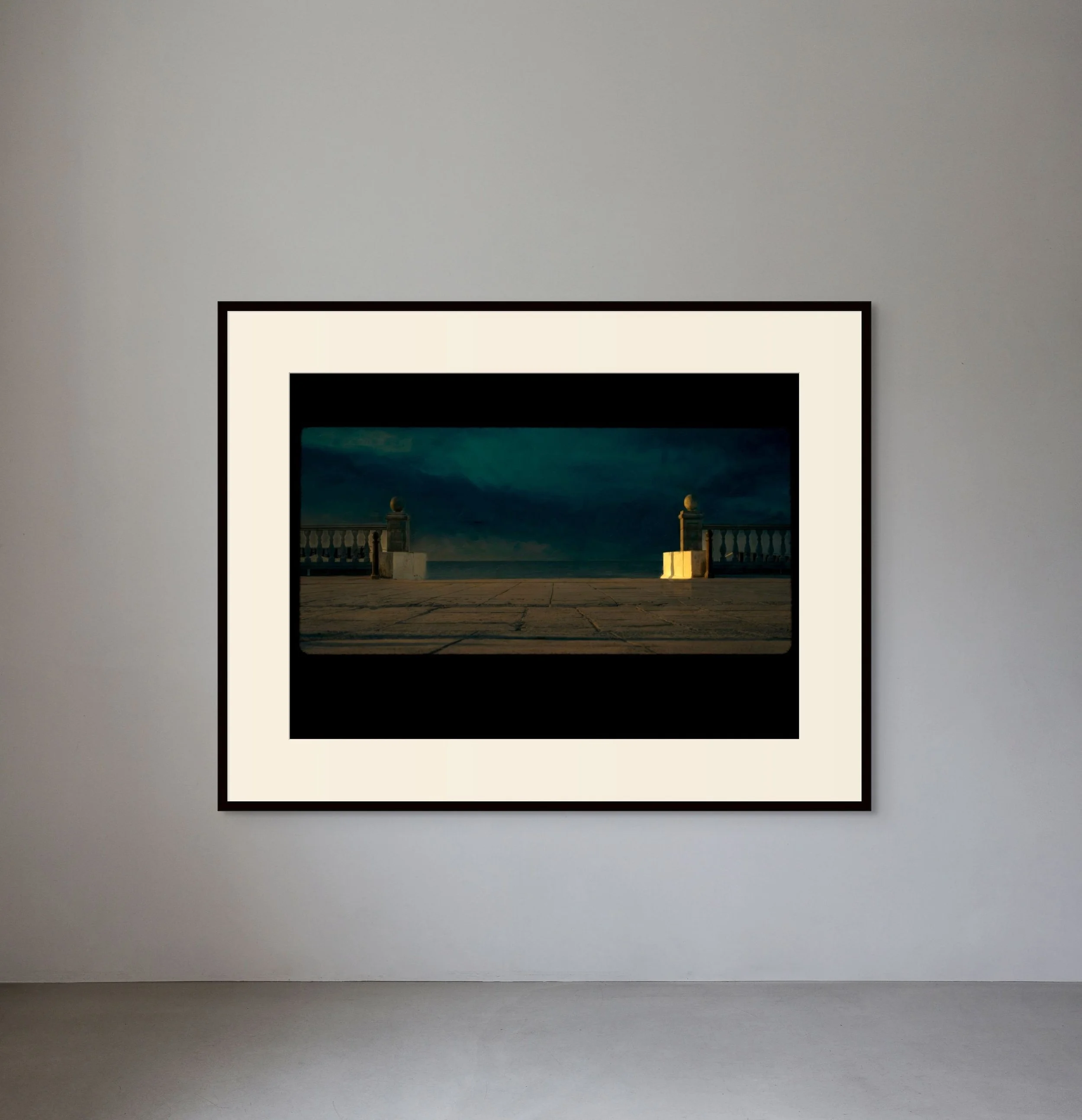 Photograph of a framed artwork on a white gallery wall showing a terrace with balustrades and pillars under a dark, cloudy sky at dusk or dawn.