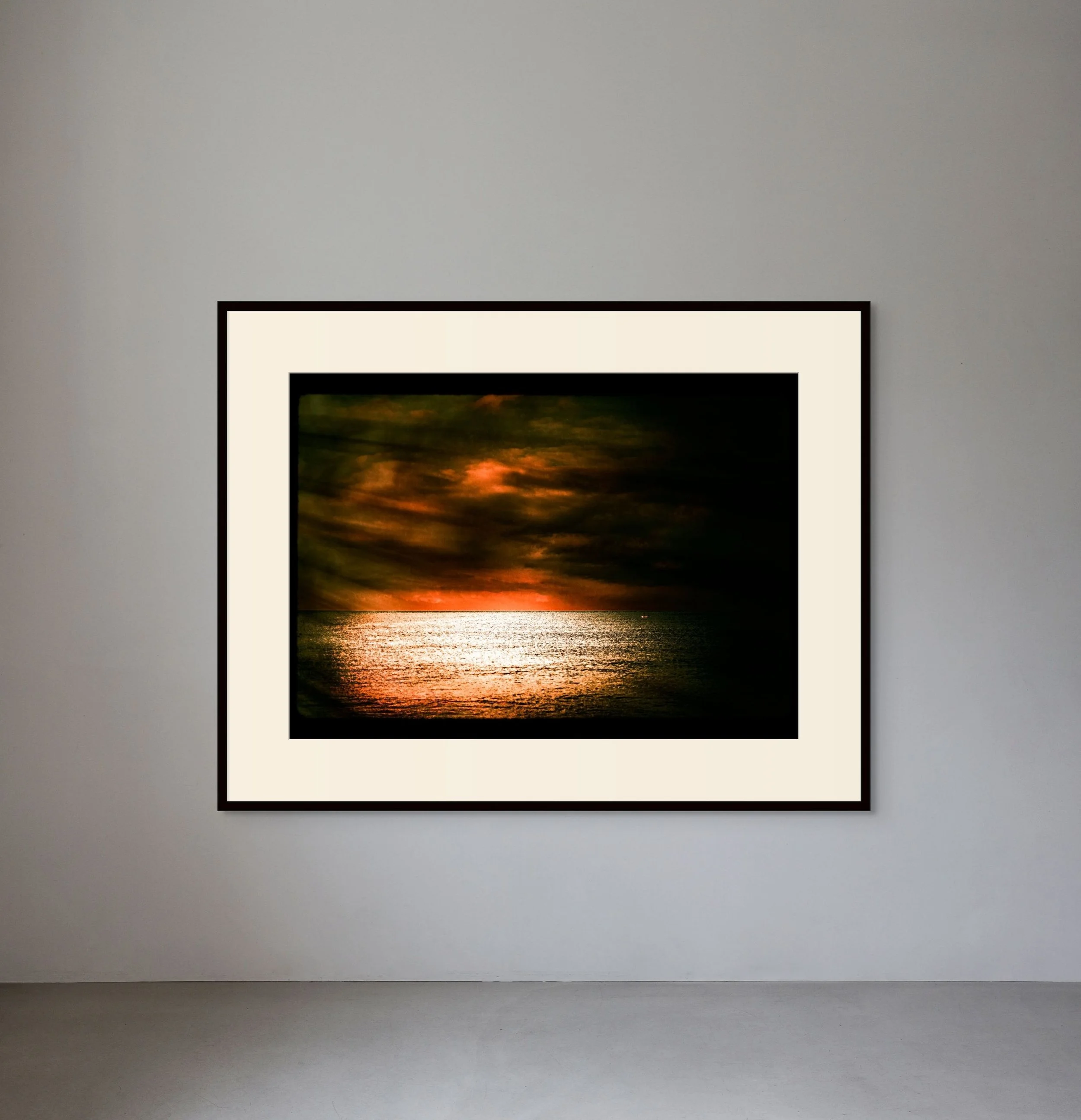 Framed sunset photograph of a dark sky with clouds over a reflective ocean, indoors against a plain wall.