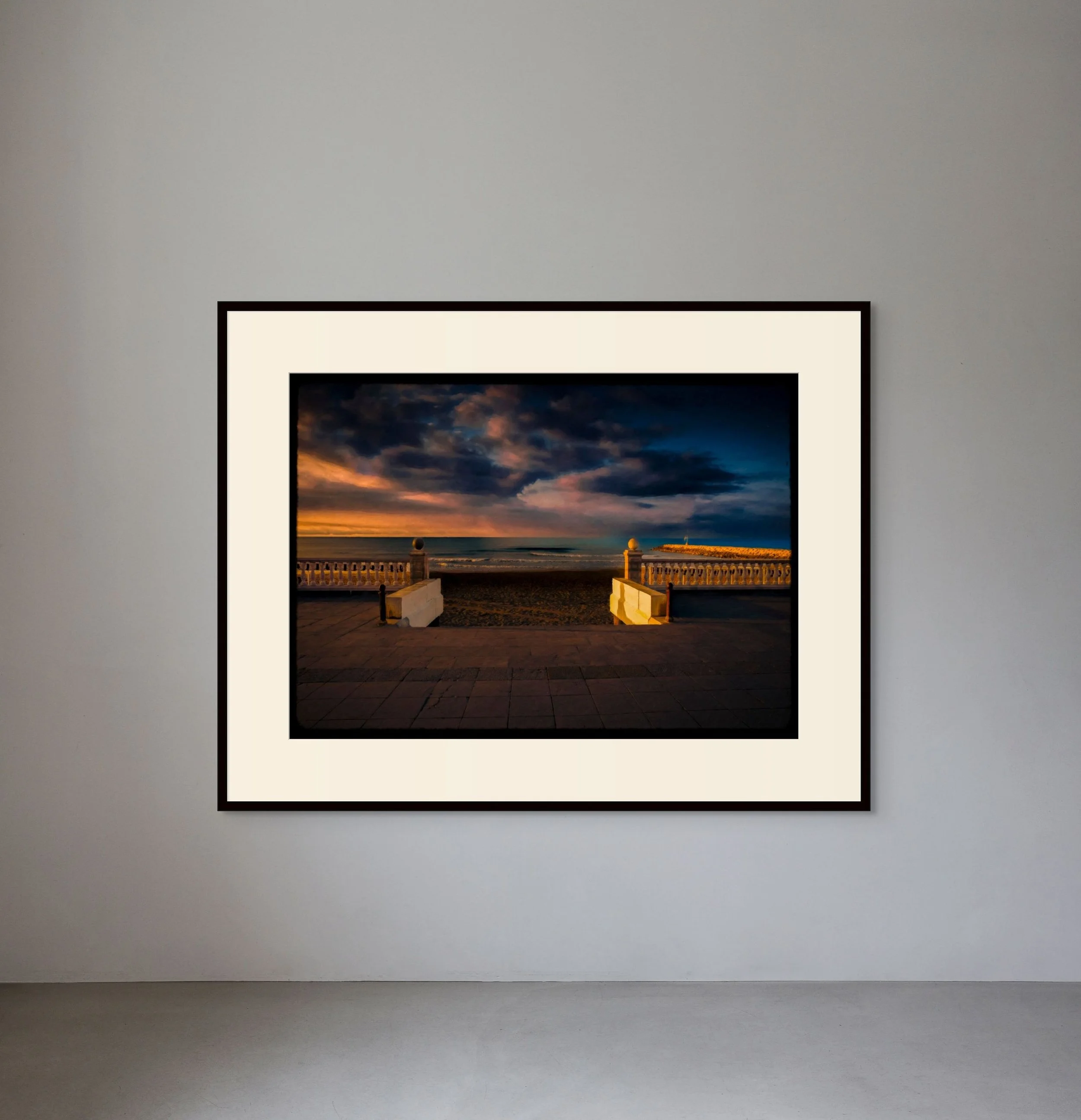 Framed photograph of a sunset over the beach with clouds, ocean, and a railing in the foreground