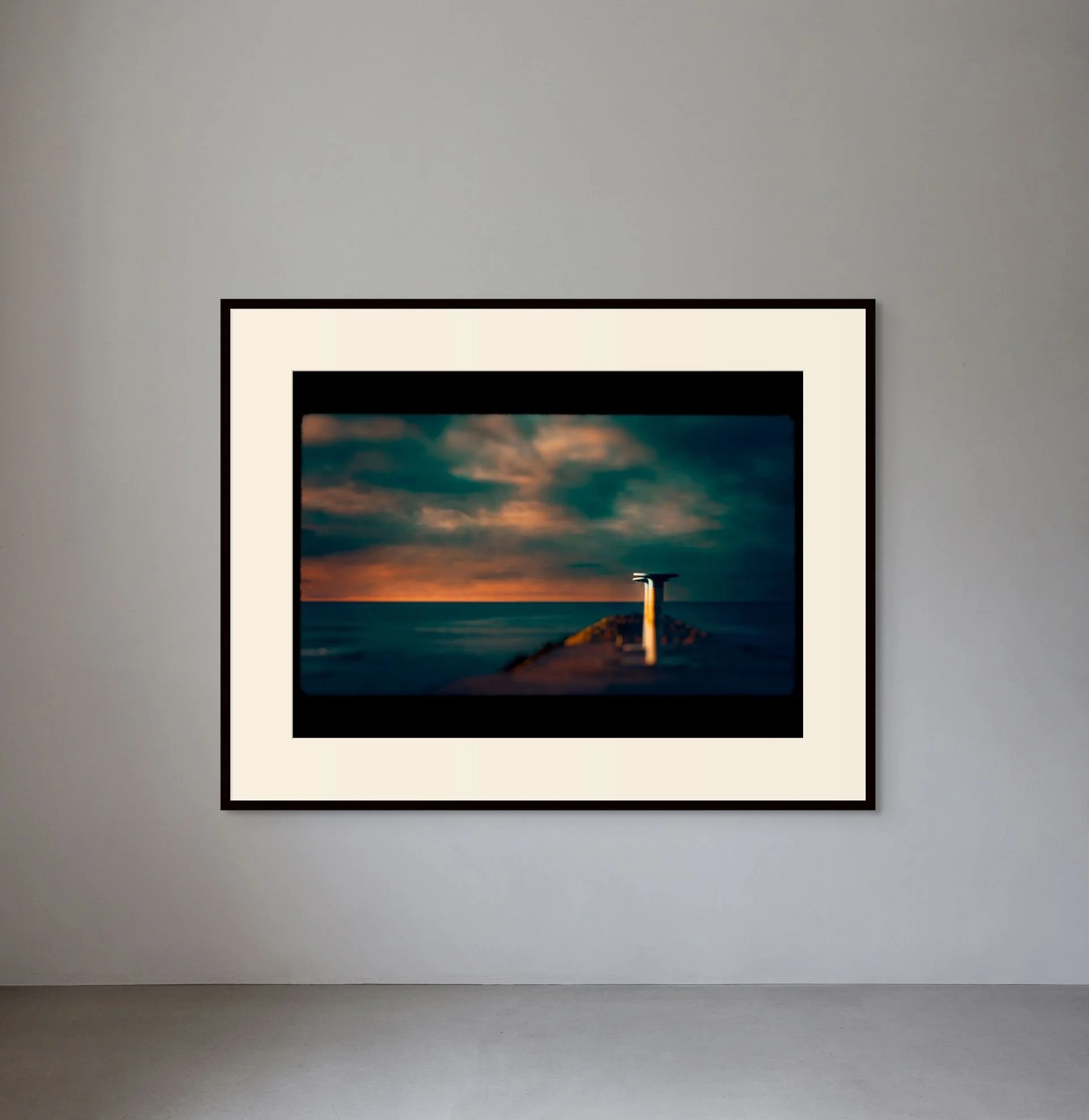 A framed landscape photograph on a plain wall showing a dock extending into a body of water during sunset, with a cloudy sky.