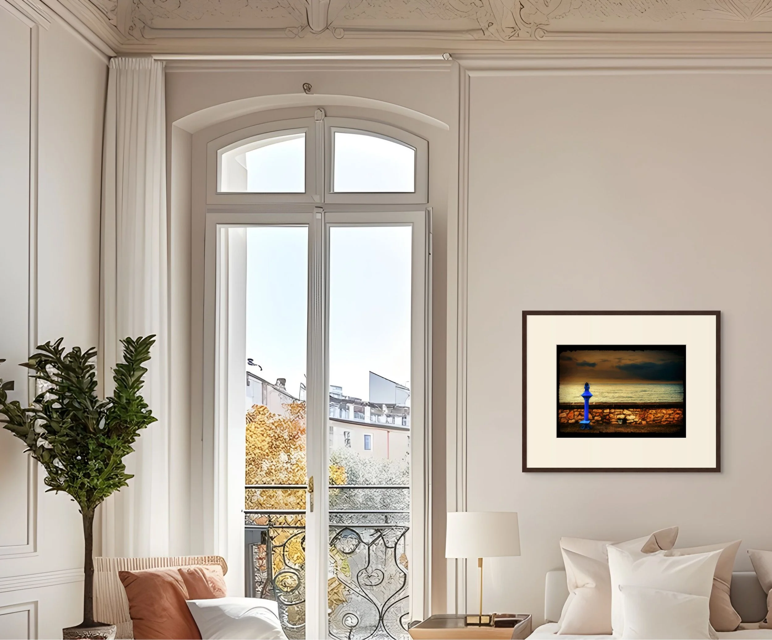 A bright living room with large arched window, white walls, a framed painting of a lighthouse at sunset, white couches with beige and brown pillows, a floor lamp, a potted tree, and a balcony with decorative iron railing, views of neighboring buildin