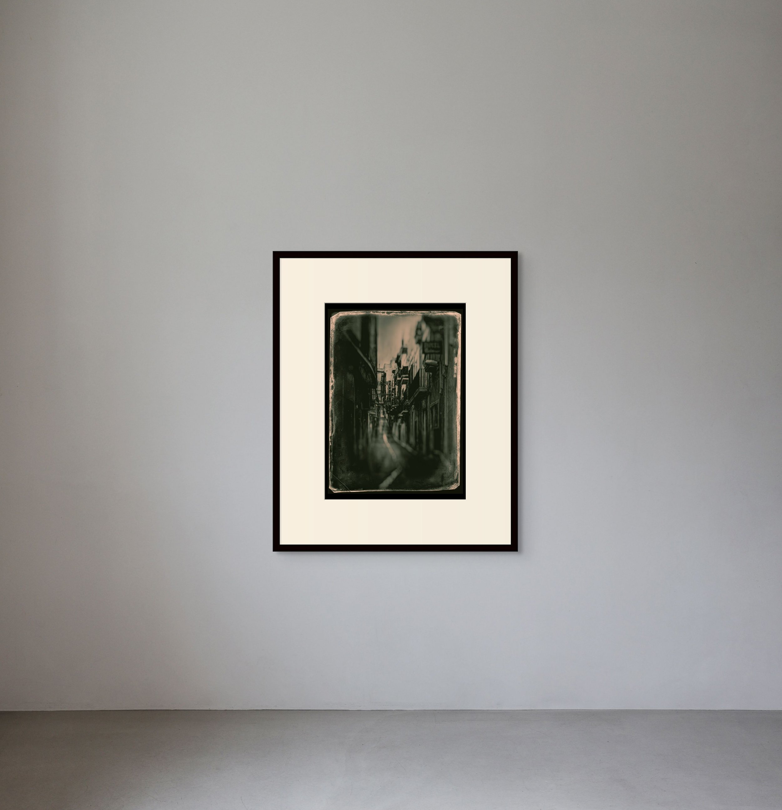 Framed black and white painting of a city street hanging on a light gray wall in an art gallery.