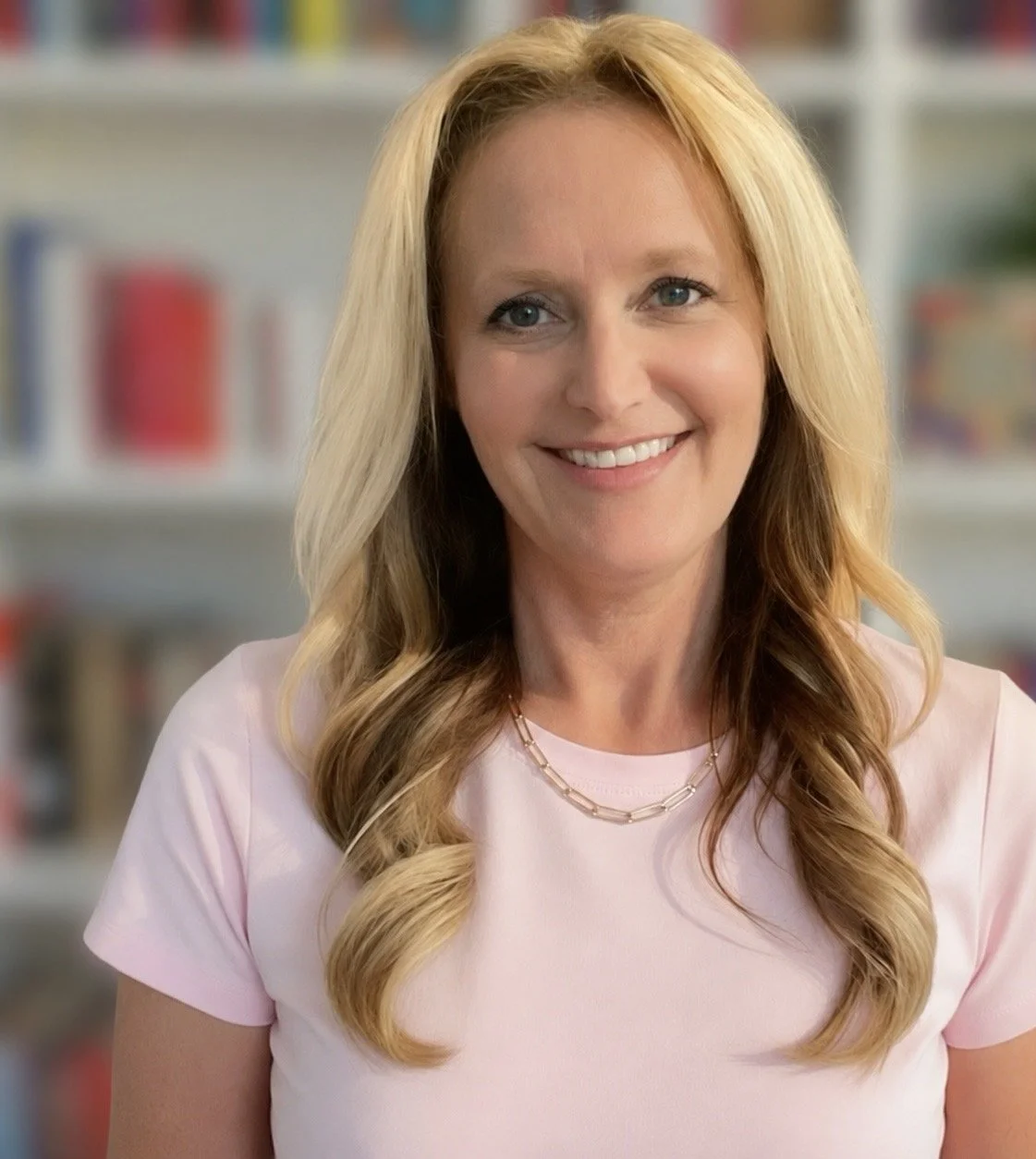 A woman with long blonde hair smiling, wearing a light pink shirt and a gold chain necklace, standing in front of a blurry bookshelf background.