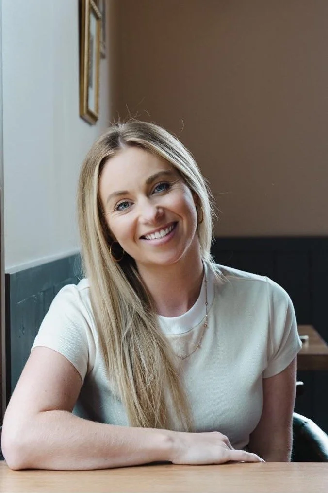Smiling woman with long blonde hair, wearing a white t-shirt, seated at a wooden table in a cozy indoor setting.