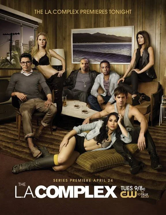 Seven young adults in a living room with wood-paneled walls, a large window, and a landscape painting. They are posed casually on and around couches, with a coffee table in the center. The text promotes the premiere of the TV series "The L.A. Complex