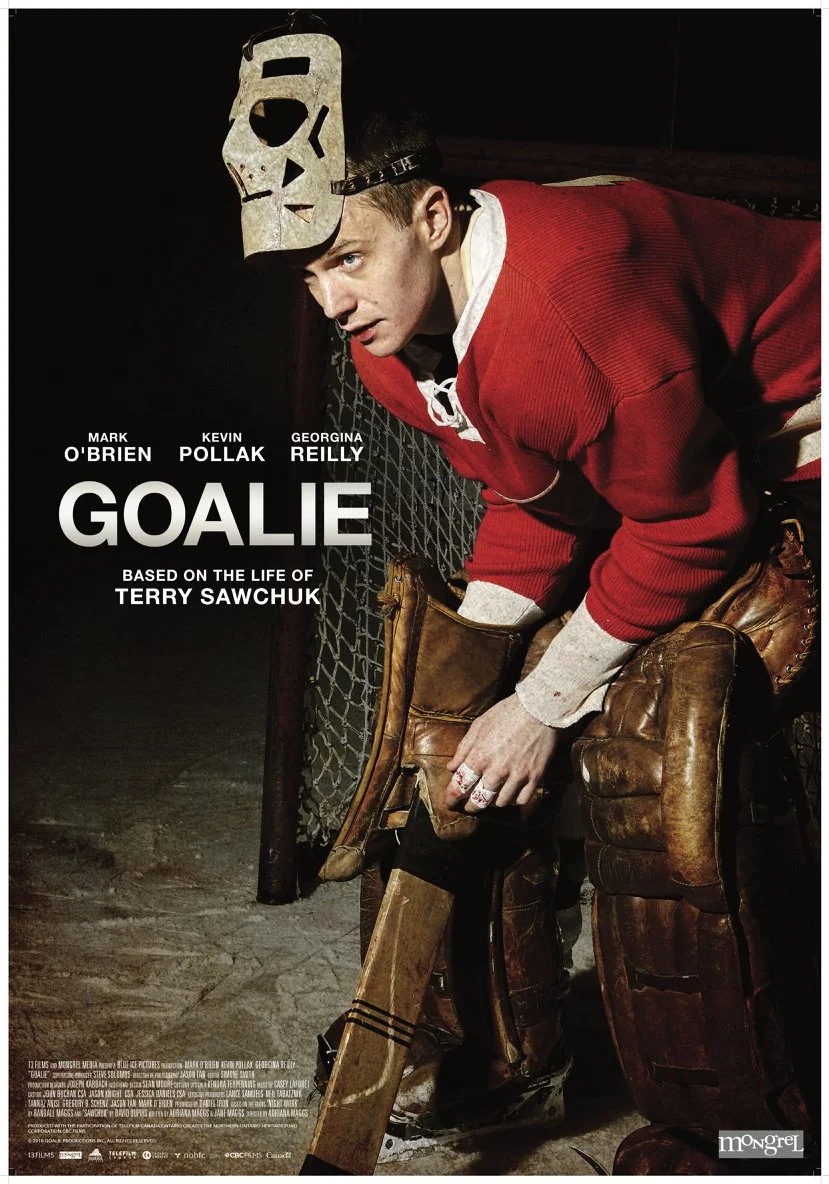A hockey goalie wearing a red jersey and brown pads, leaning forward with a serious expression, holding a hockey stick, with a mask hanging on his head, in a dimly lit setting.