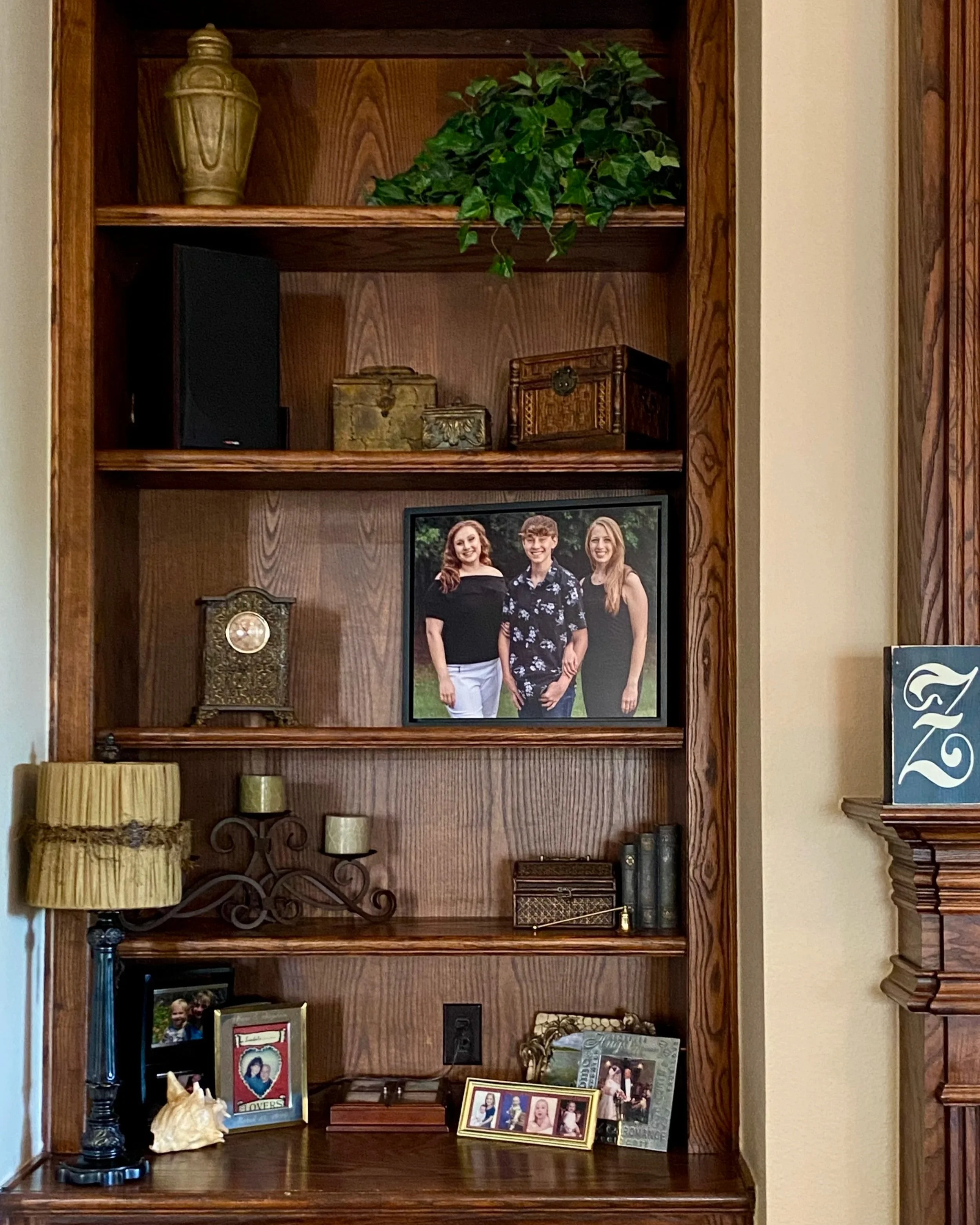 Wooden bookshelf with decorative items, a family photo in the center, framed photos and personal objects on the bottom shelf, and a potted plant on the top shelf.