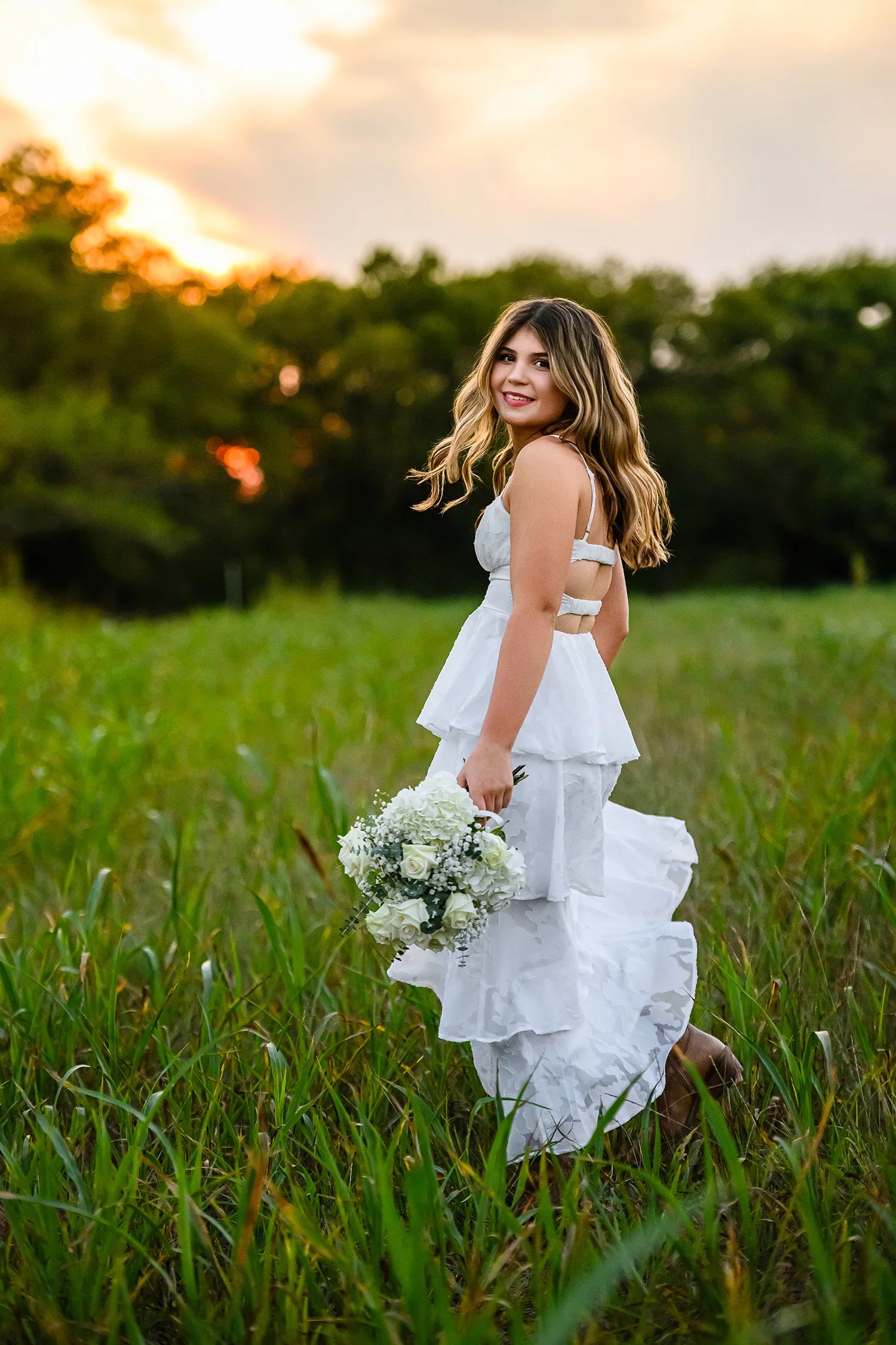 A young woman in a white dress and cowboy boots looks back at camera while running in a green field at sunset
