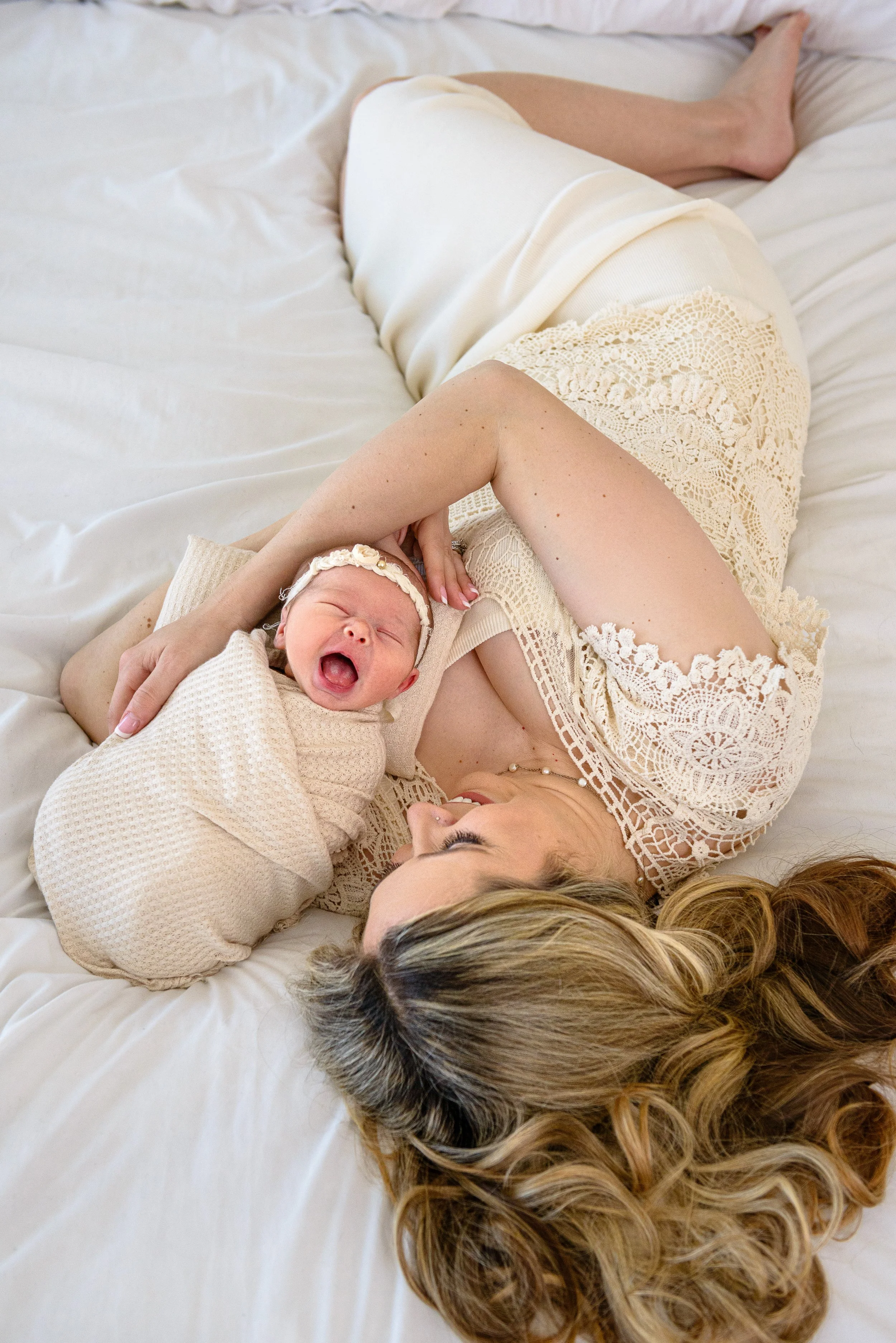 A mom smiles while holding her yawning newborn baby girl on a bed