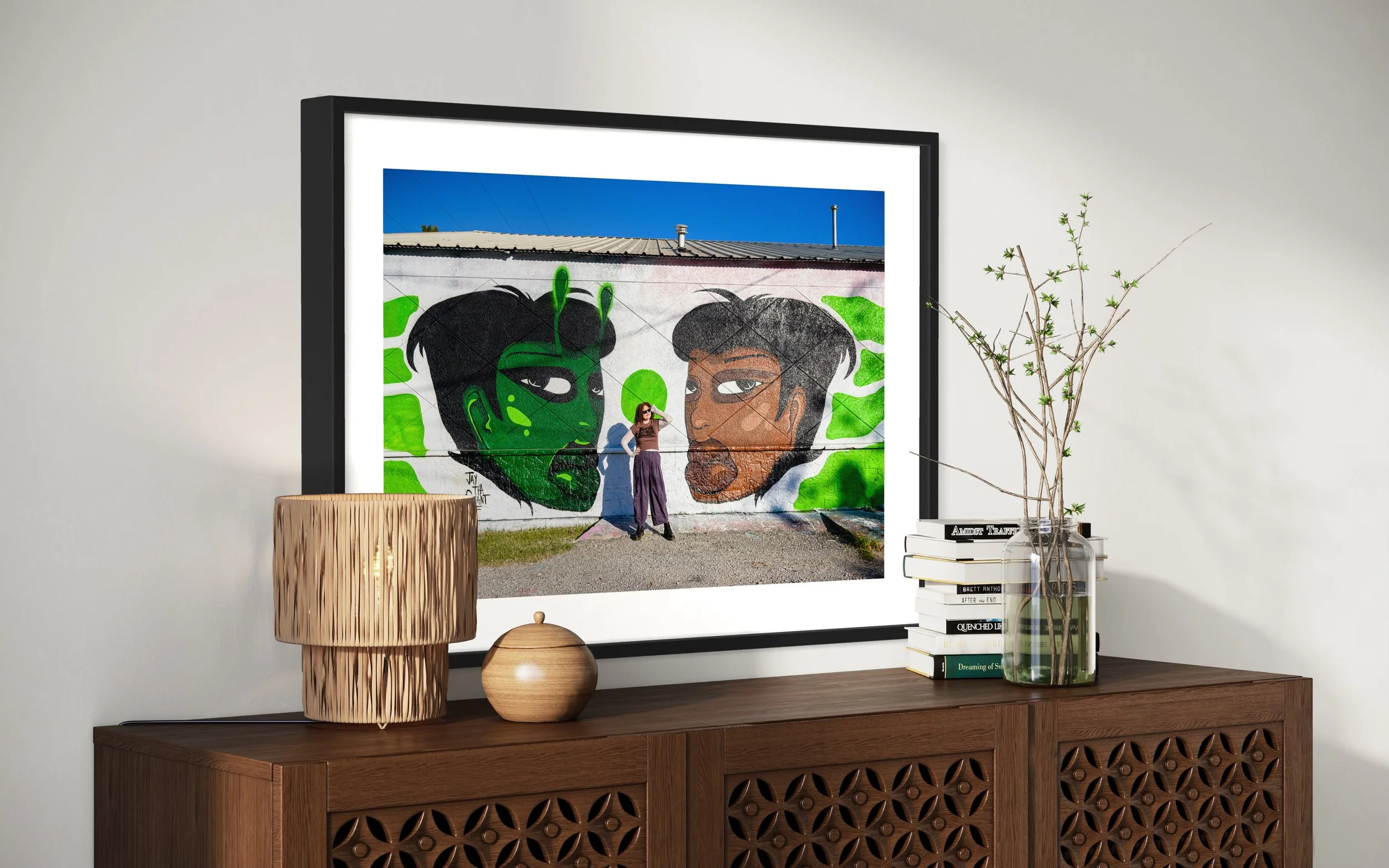 Photo of a wall art mural depicting two characters with stylized faces, one with dark hair and makeup, and the other with short hair and a beard, painted on an outside wall with green accents.