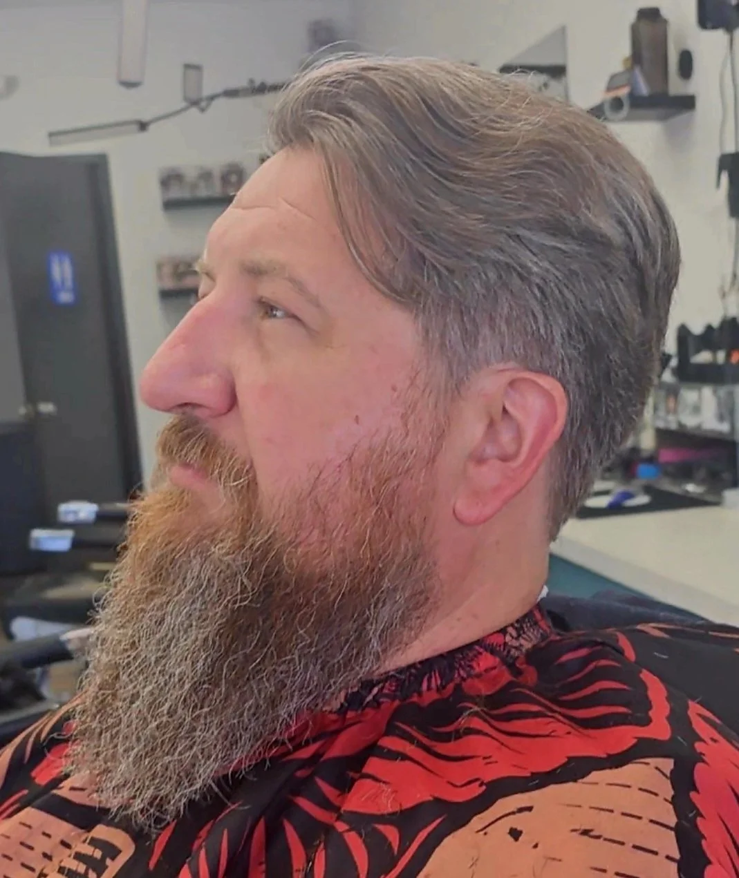 Side profile of a man with a full beard and slightly wavy hair, sitting in a barber shop.