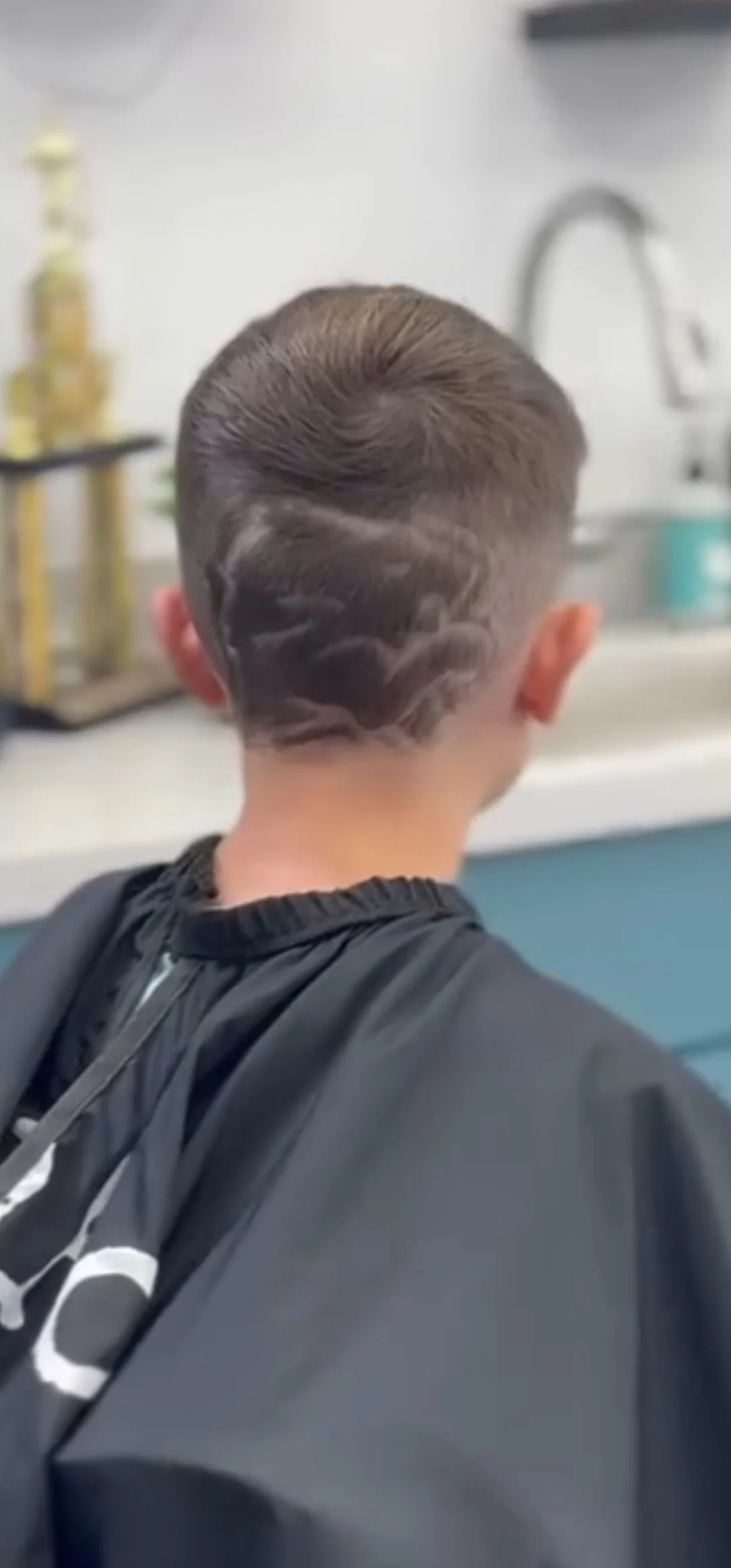Back view of a person with a short hairstyle featuring a design shaved into the hair, wearing a black jacket, in a salon or barbershop setting.