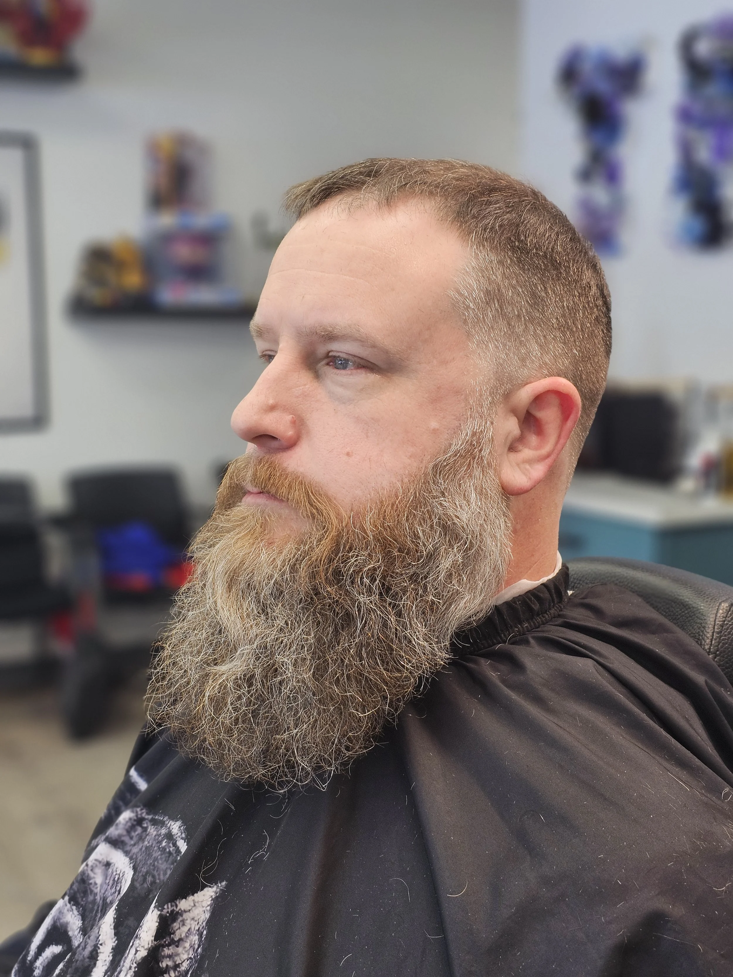 Close-up of a man with a full beard and short brown hair getting a haircut in a barbershop.