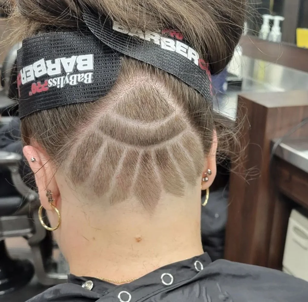 Back of a person's head with an intricate shaved hairstyle featuring geometric patterns, wearing a black and red baseball cap and multiple earrings.