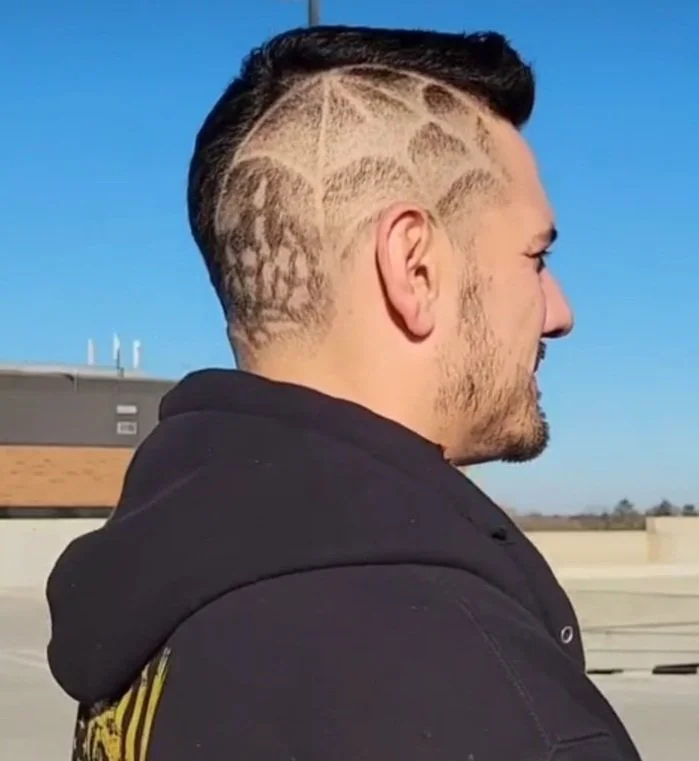 Side view of a man with a hairstyle featuring shaved sides and designs, wearing a black hoodie, standing outdoors against a clear blue sky.