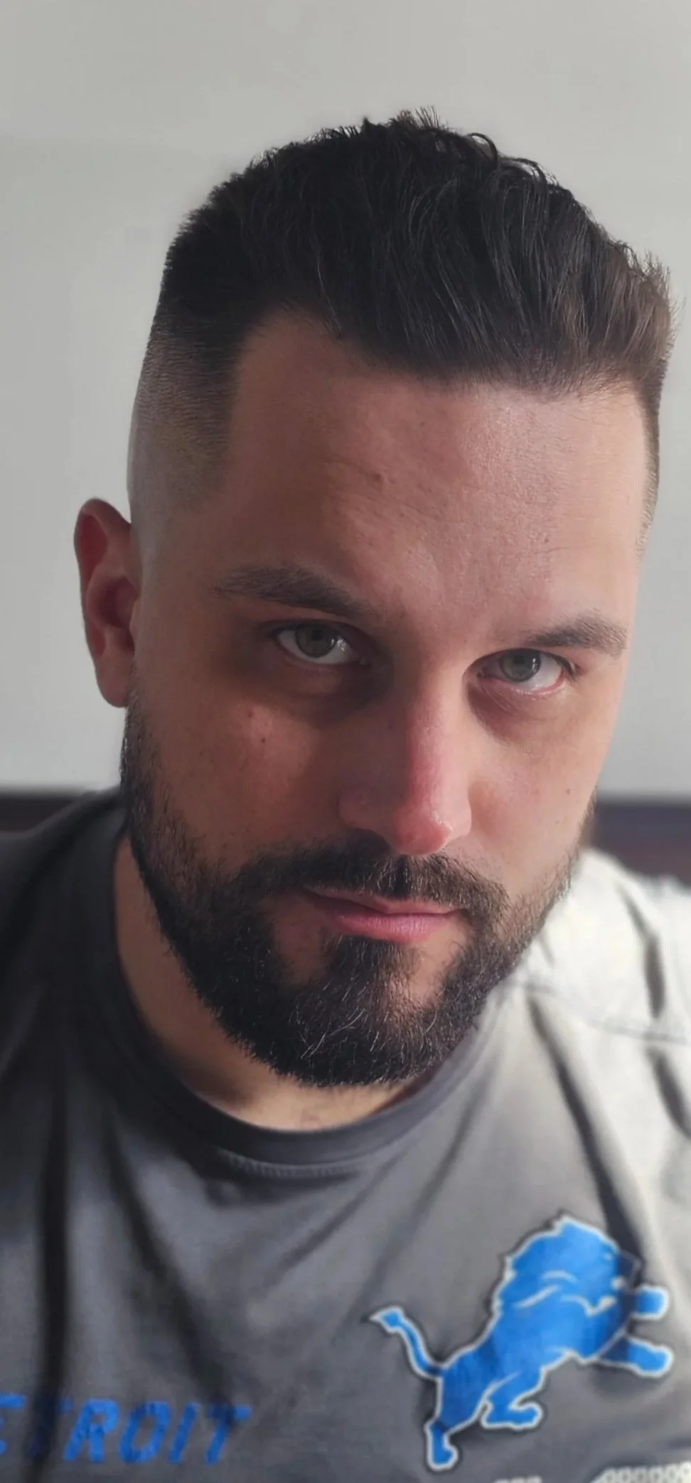 Close-up of a man with dark hair and beard wearing a gray Detroit Lions shirt.