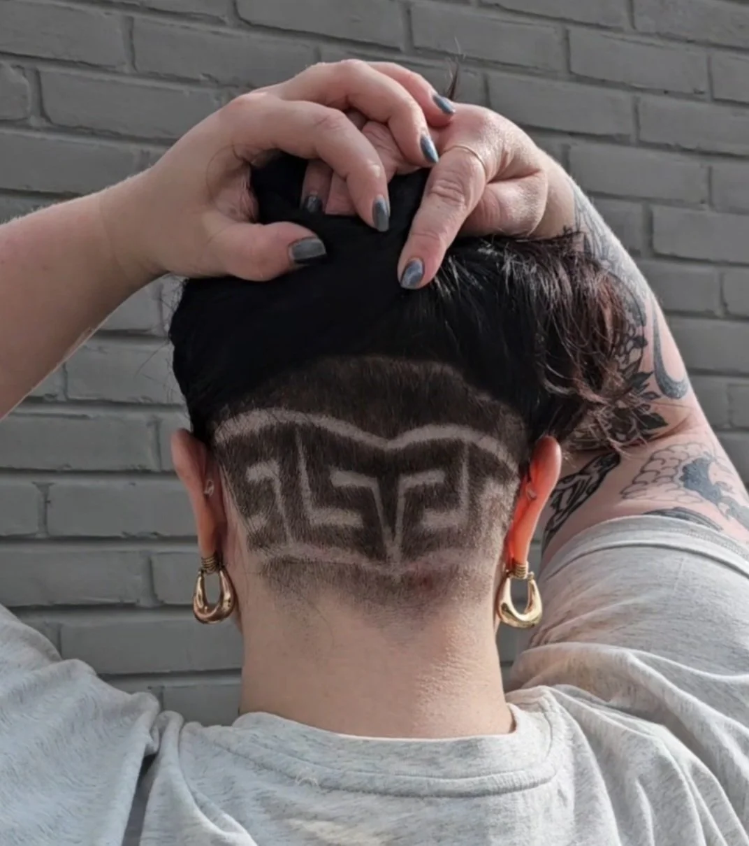 Back of a person's head with a hair design shaved into the hair, spelling the word 'GIRL.' The person has tattoos and wears large hoop earrings, with grey and black nail polish.