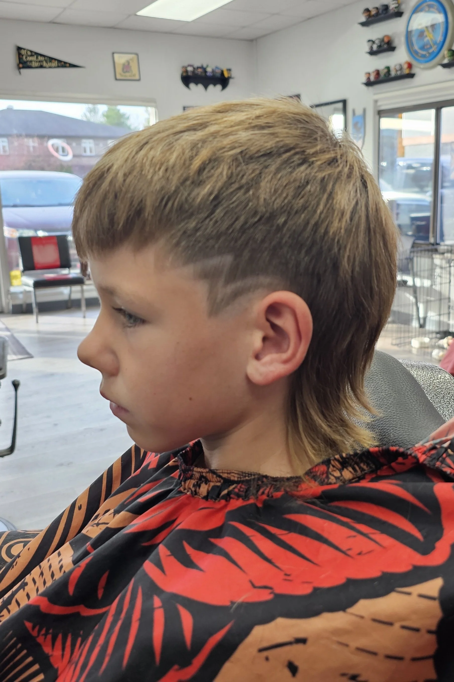 A young boy with light brown and blonde hair sitting in a hair salon chair with a black and red patterned cape around his shoulders. The salon interior has white walls, shelves with toys, and a large window showing parked cars outside.