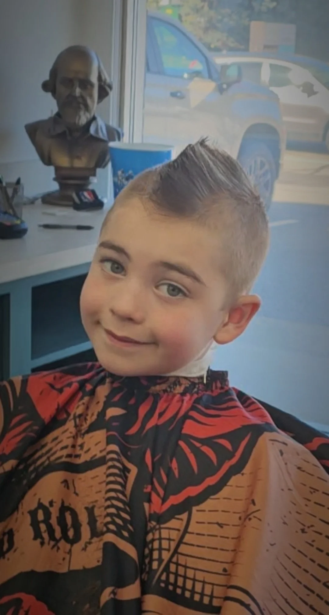 Young boy with short hair and a vibrant patterned shirt taking a selfie inside a barber shop. A bust sculpture of a man is visible on the counter behind him, along with a blue cup and a car visible through the window.