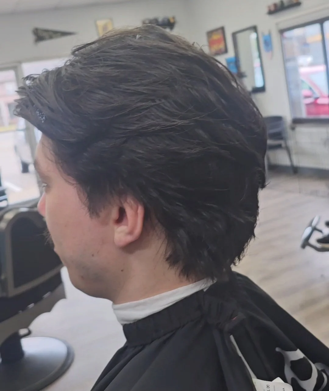 Side view of a man with dark, styled hair sitting in a barbershop chair, wearing a black barber cape inside a barbershop