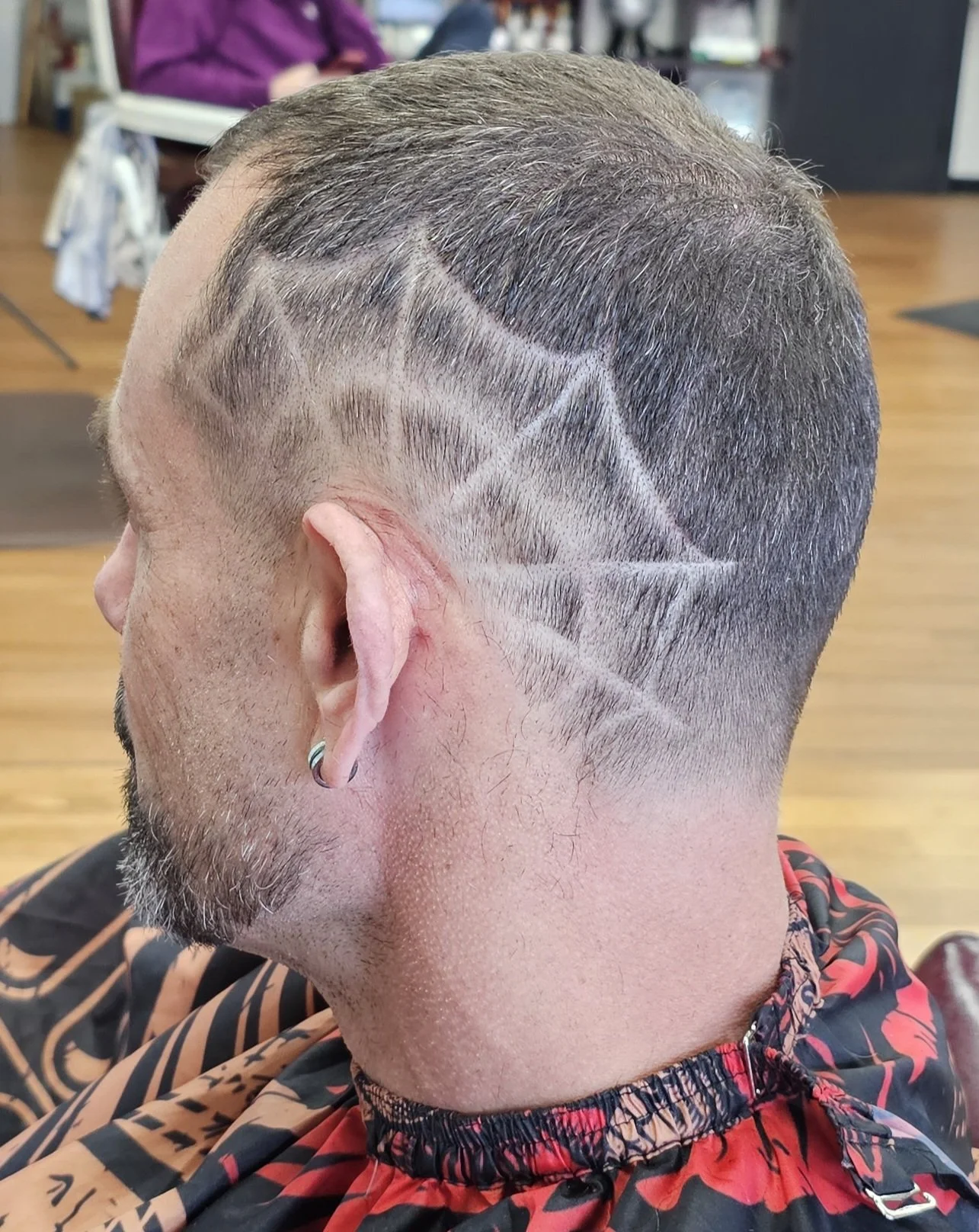 Man with a short haircut with geometric shaved design on side of head, wearing a red and black patterned cape in a barbershop.