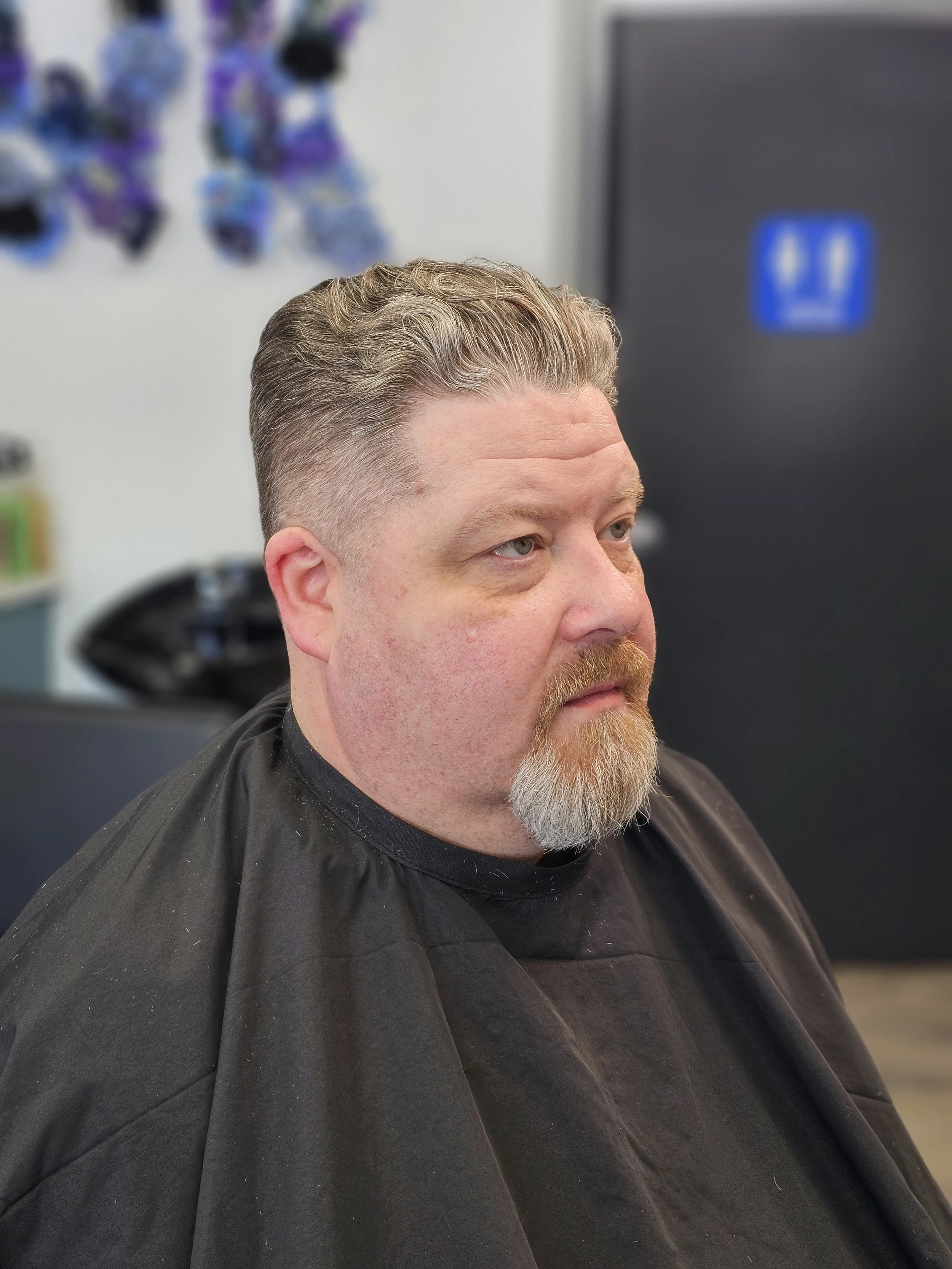 Man with blonde hair and a beard sitting in a barber's chair, wearing a black salon cape, in a hair salon.