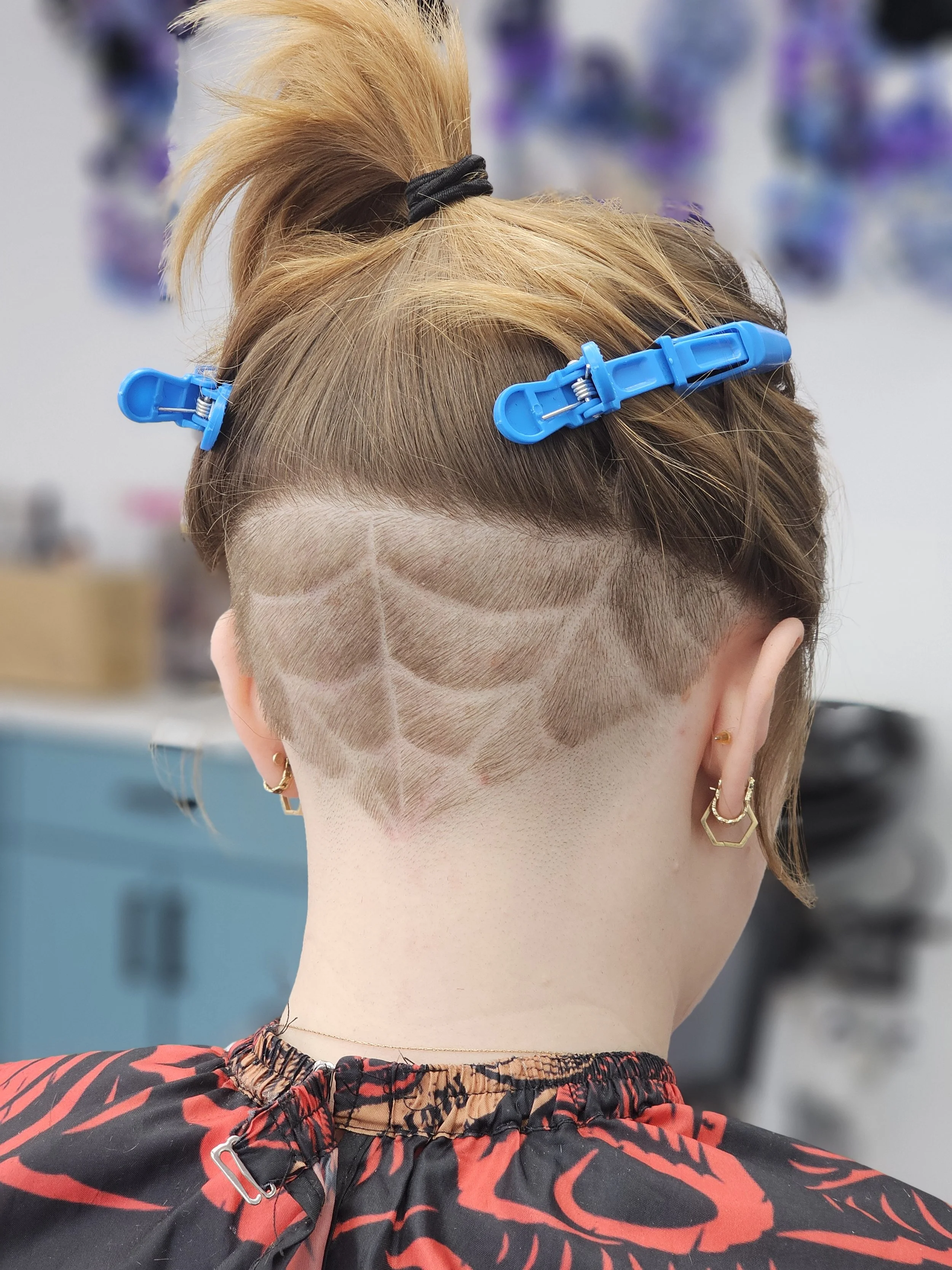 Back of a person's head with short hair, partially shaved with a patterned design, and a top section tied up with hair clips. The person is wearing earrings and a patterned shirt or jacket.