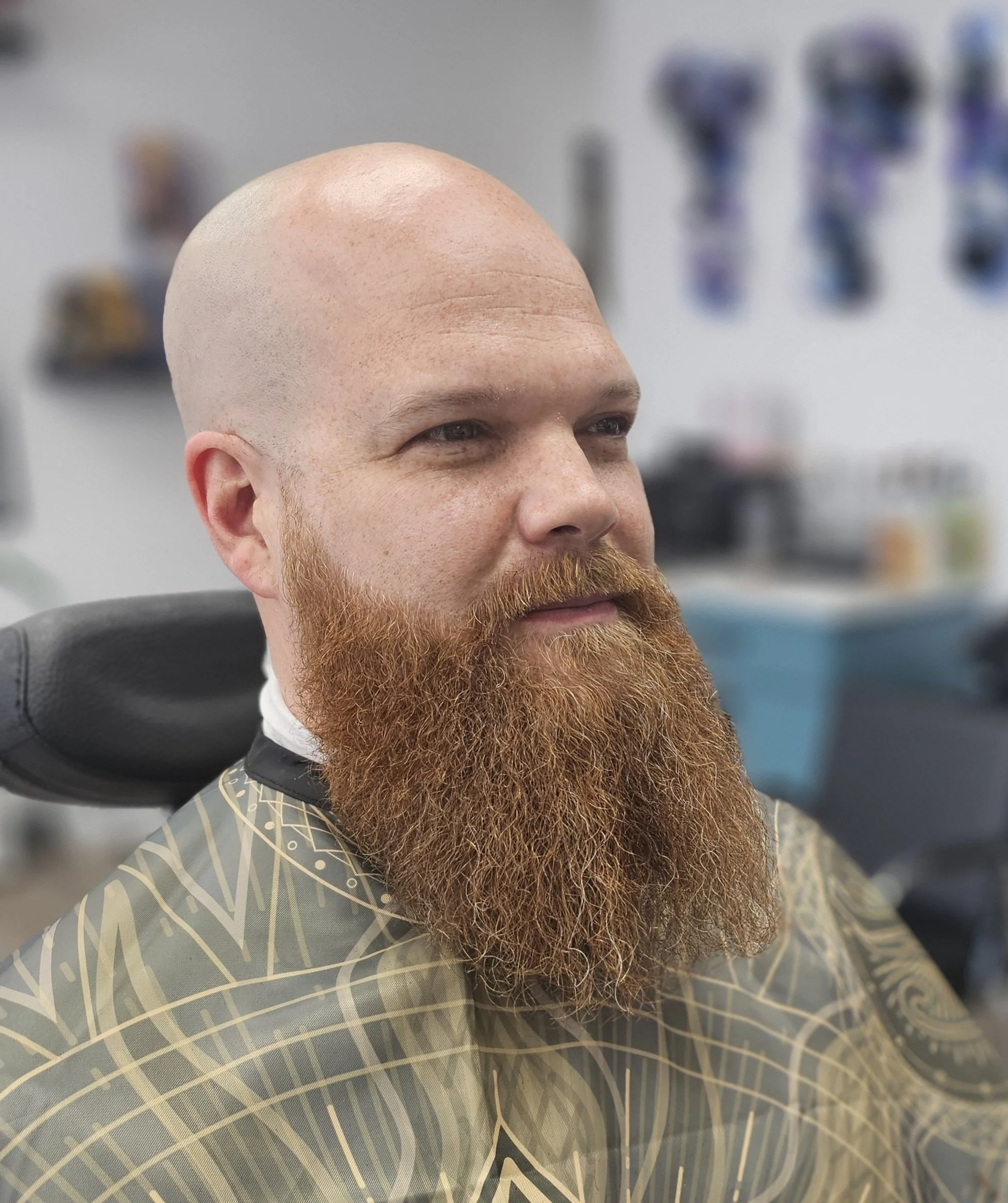 Close-up of a man with a bald head and a full reddish beard, sitting in a barber shop chair, with barber tools blurred in the background.