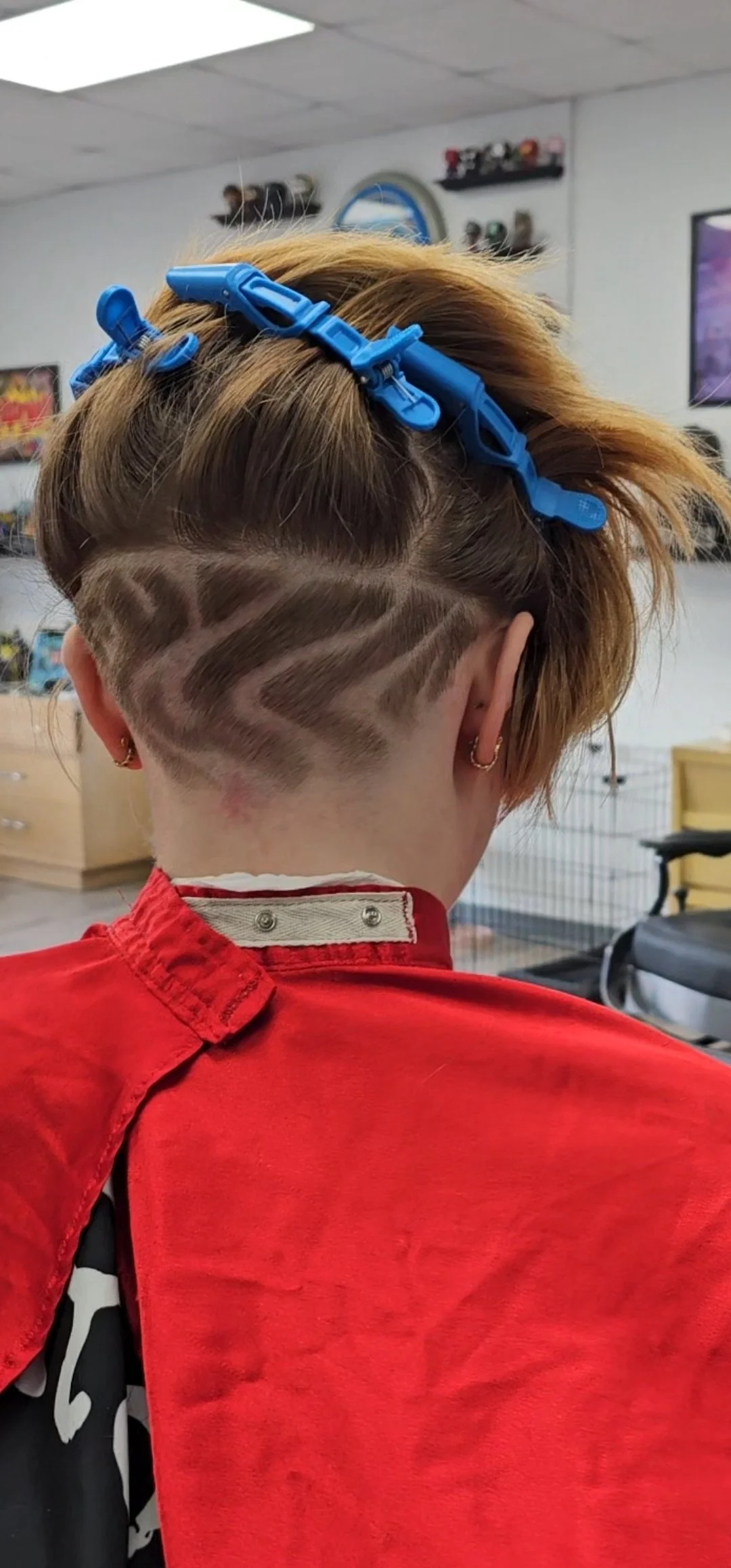 Back of a person's head with a stylized haircut and shaved pattern, wearing a red barber cape and a blue hair clip.