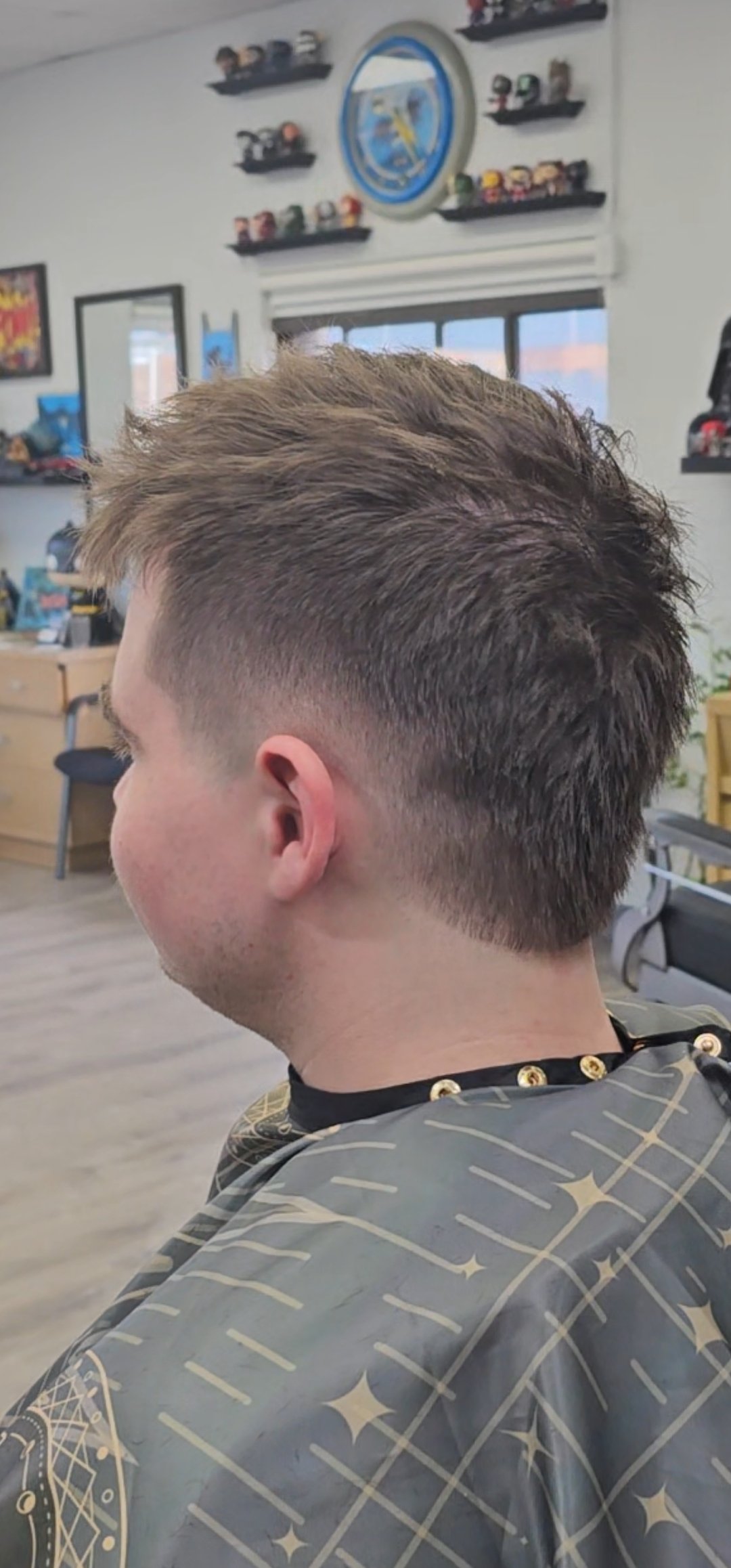 Side view of a man with a modern, textured haircut featuring a fade on the sides and longer, styled hair on top, inside a barber shop with framed pictures, a clock, and shelves on the wall.