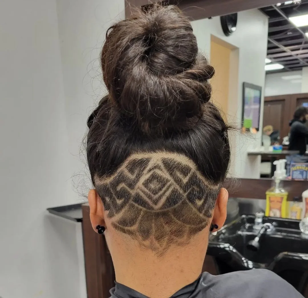 Back of a person's head with an elaborate hairstyle and a geometric shaved tattoo pattern in the hair.