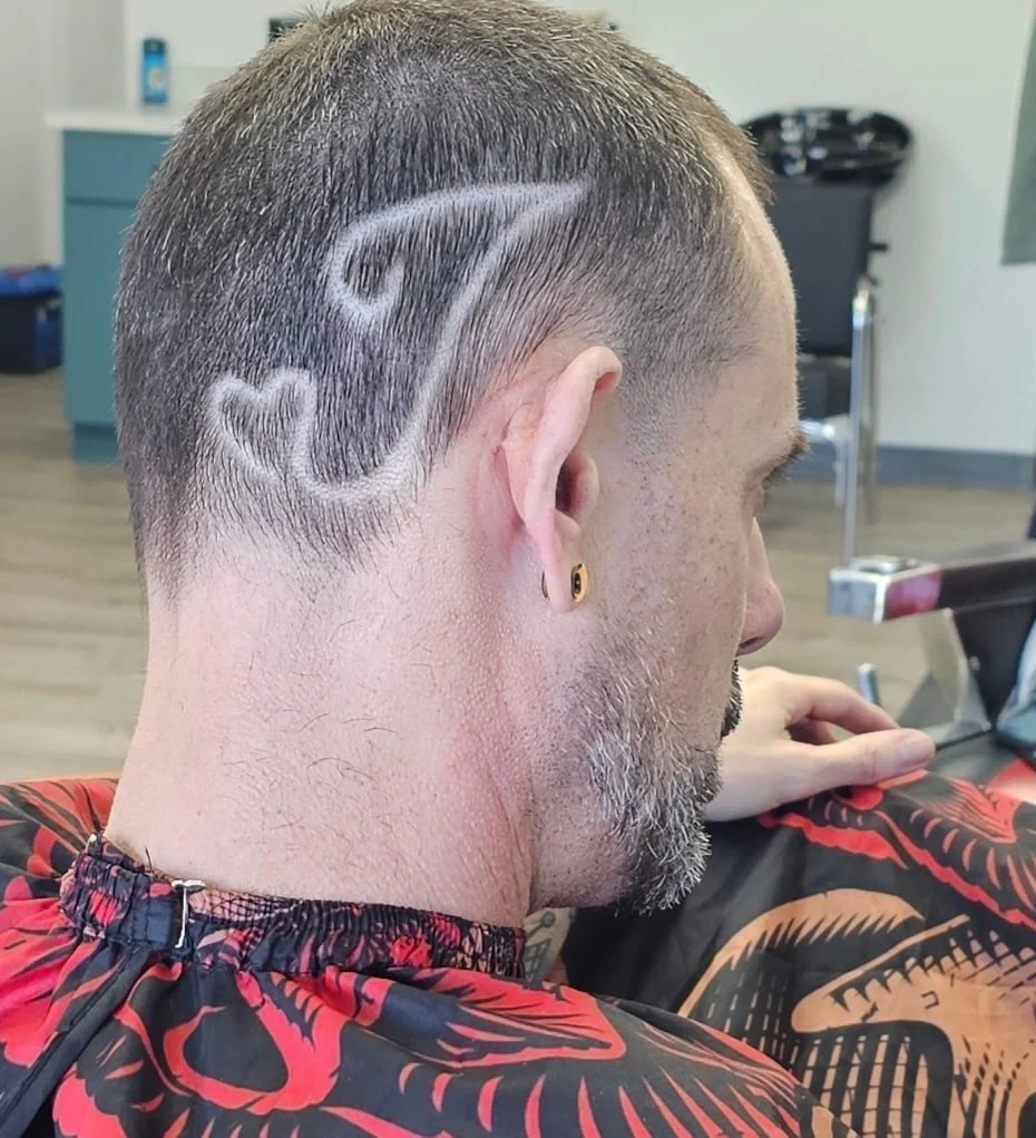 A man at a barber shop has a haircut with a design shaved into the side of his head, featuring a stylized letter 'G' and a heart shape.