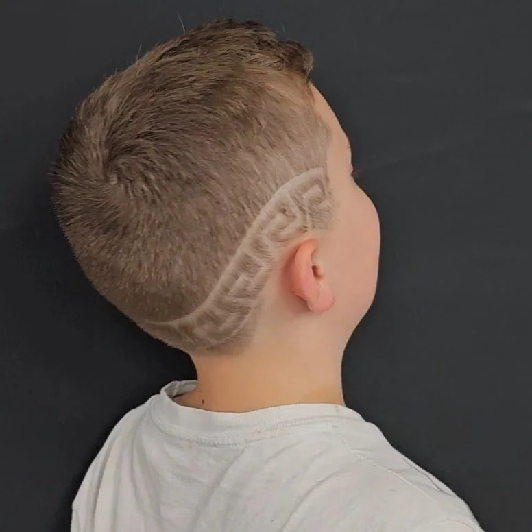 Side view of a boy with short blonde hair featuring a shaved design pattern on the back of his head.