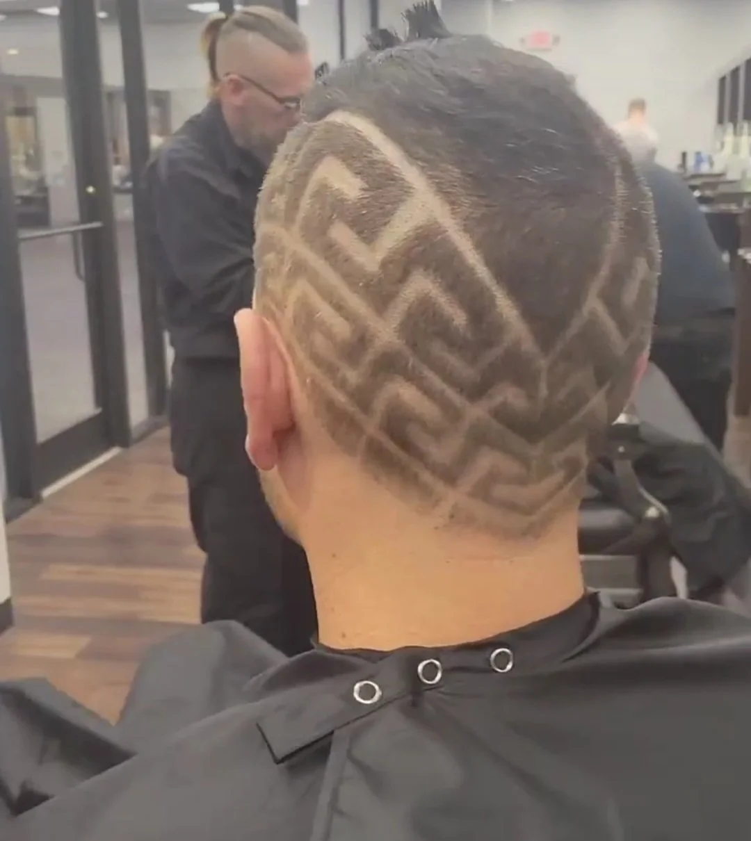 A person with a shaved head featuring a geometric pattern haircut in a salon, with a hairstylist working in the background.