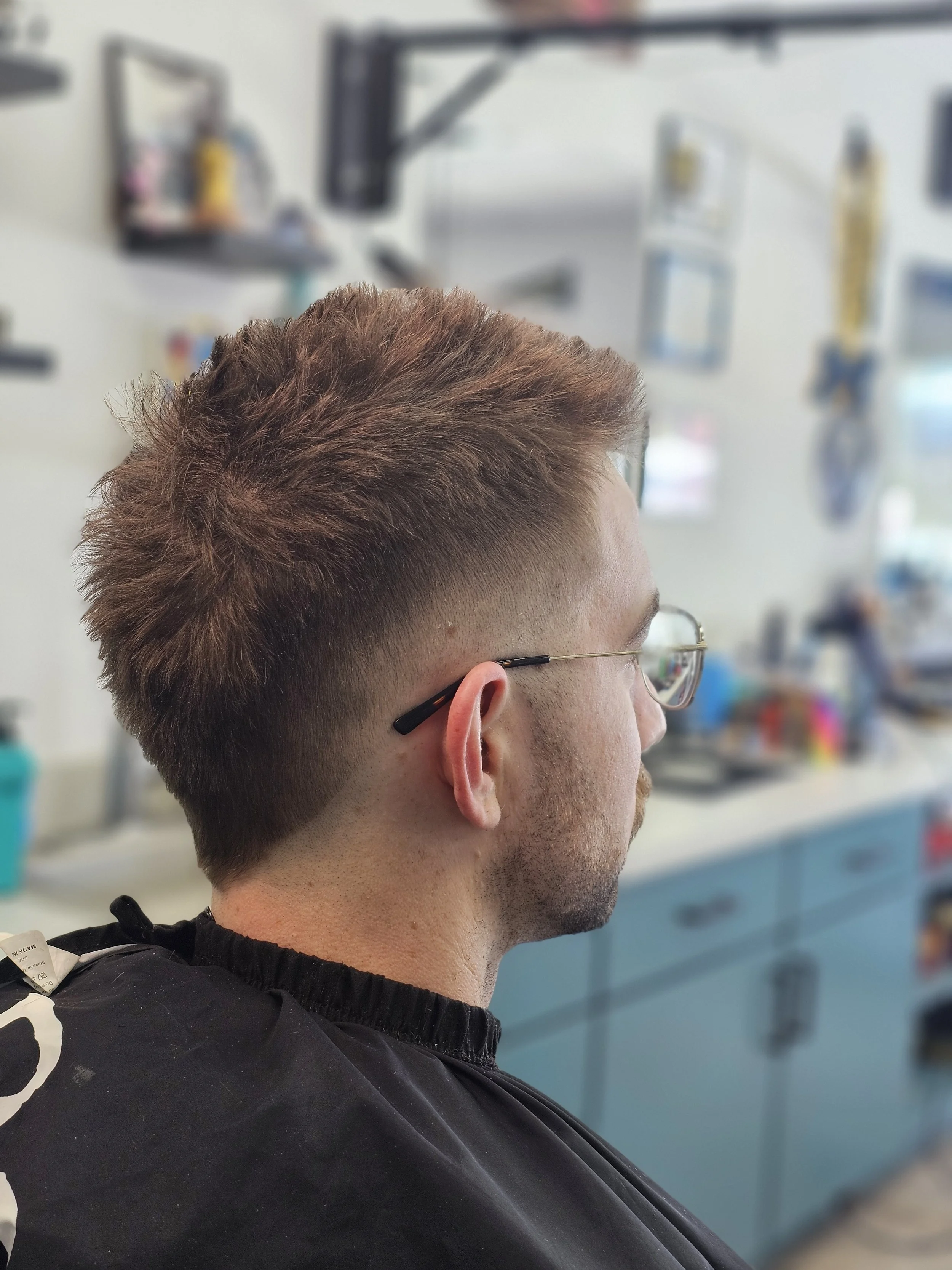 Side view of a man with a fresh men’s haircut, short on the sides and back, longer on top, wearing glasses, in a barbershop setting.