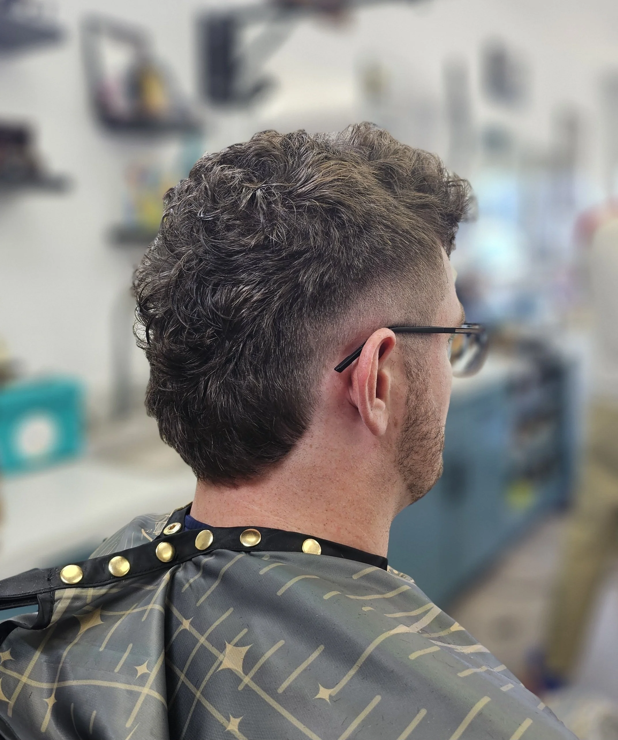 Close-up of a person's head with short, curly, brown hair and glasses, wearing a barber cape with gold buttons, in a barbershop.