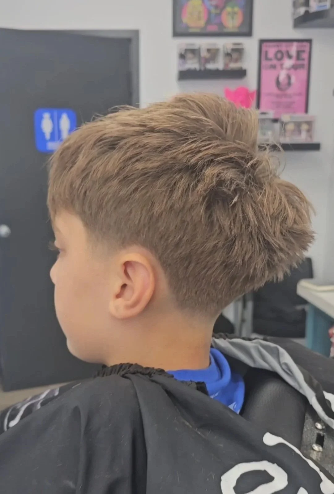 Side view of a young boy with a short, layered haircut in a barbershop, wearing a black barber cape with white designs, in front of a wall with posters and photos.